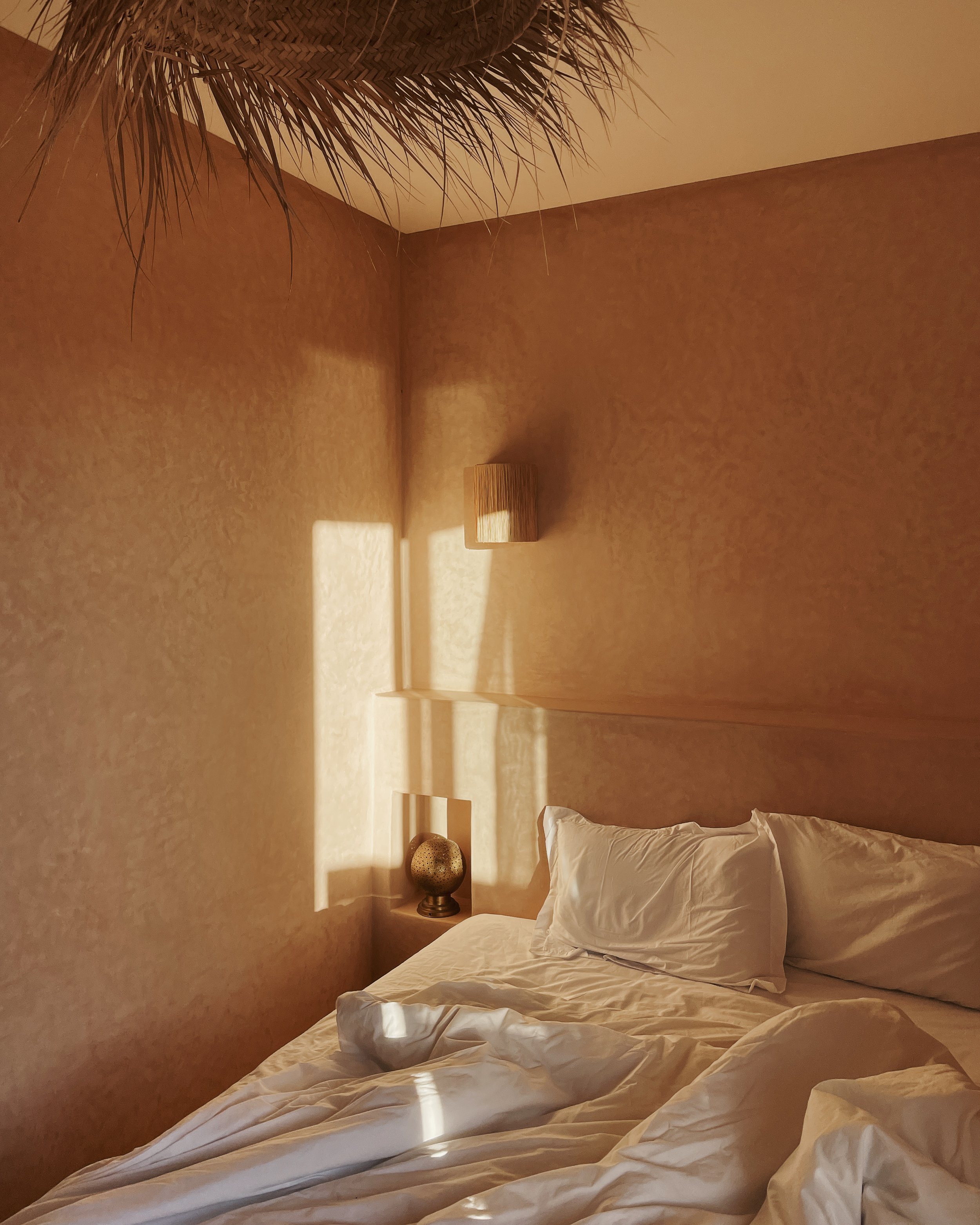 Chambre avec mur beige, lit avec draps blancs, oreillers et une petite table de chevet avec une lampe et une décoration en forme de sphère.