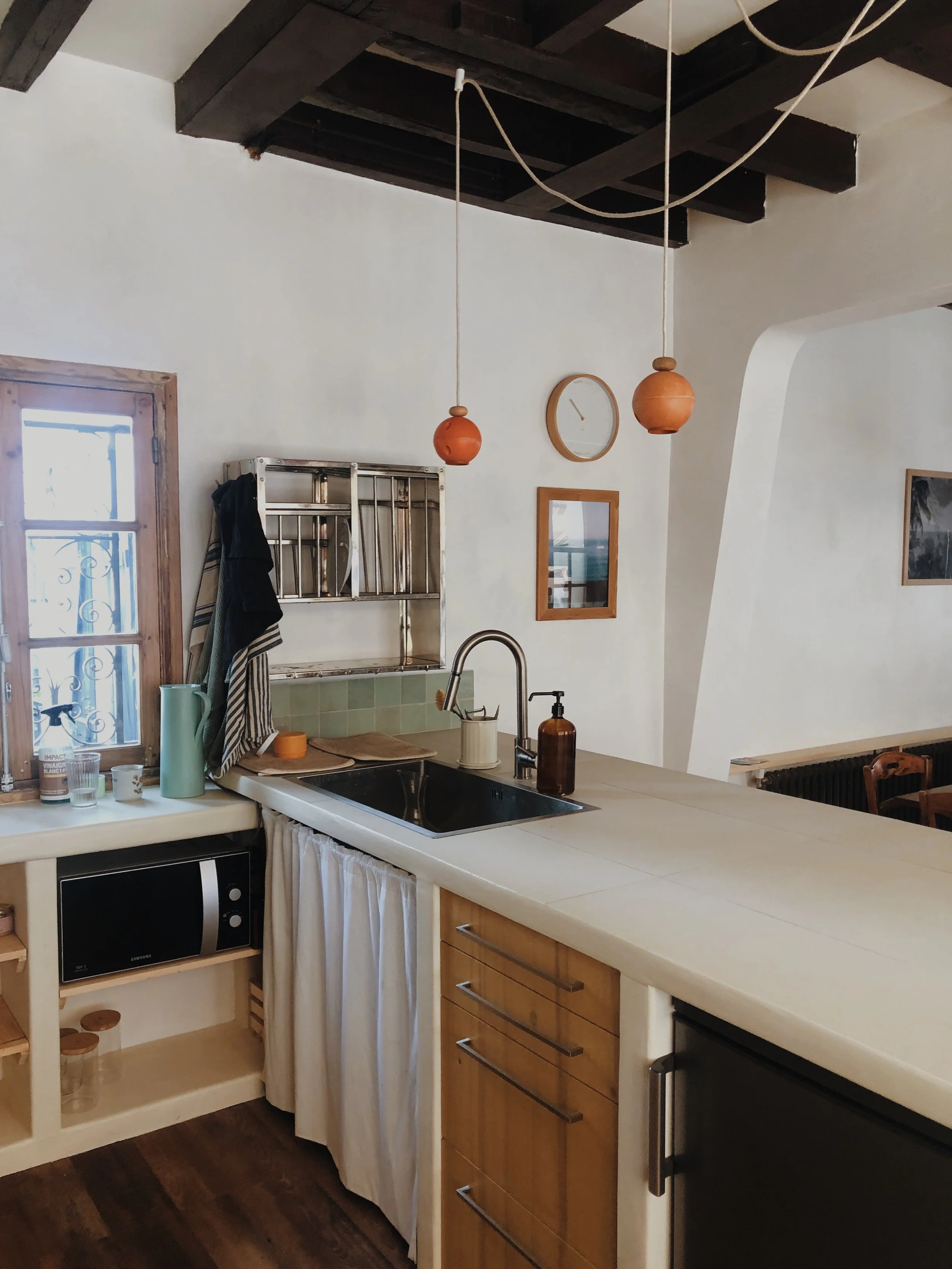 Une cuisine rustique avec un plan de travail en bois clair, un évier en acier inoxydable, un meuble à micro-ondes noir, une étagère en métal au-dessus de l'évier, et des objets comme un savon liquide, une bouteille en verre, et des tasses. Une fenêtre en bois laisse entrer la lumière naturelle, et l'ambiance est chaleureuse avec des murs blancs et un plafond en bois noir avec poutres.