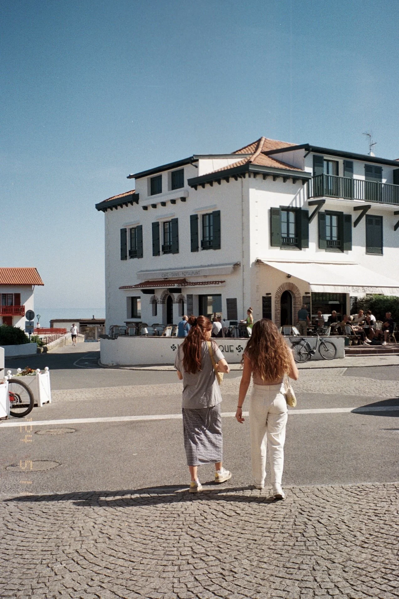 Deux jeunes femmes avec des cheveux longs marchant dans une ville ensoleillée, devant un bâtiment blanc avec des fenêtres à volets verts et une terrasse avec des tables, un restaurant avec enseigne et clientèle assise dehors.
