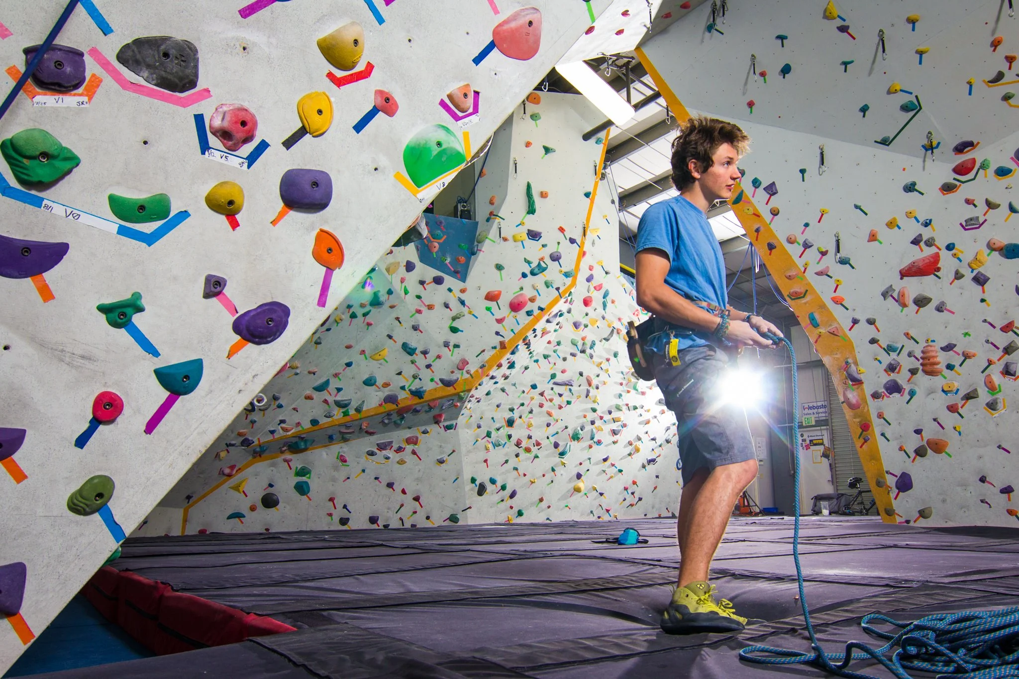 A young man in a blue t-shirt and gray shorts is belaying a climber at an indoor rock climbing gym. The climbing wall is covered with colorful holds and shapes, and there are more holds on adjacent walls. The gym has a high ceiling with natural light
