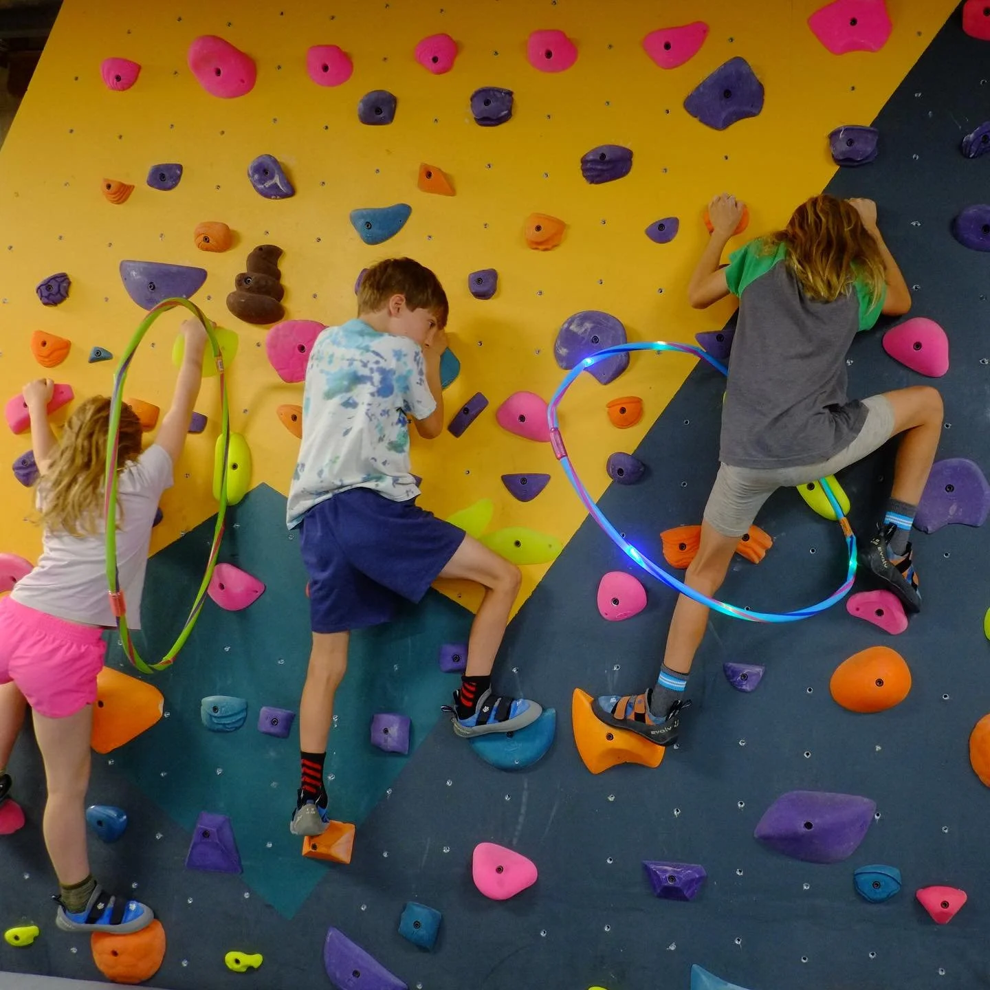 Three children rock climbing on an indoor bouldering wall with colorful holds, some of which have lights, wearing climbing shoes and harnesses.