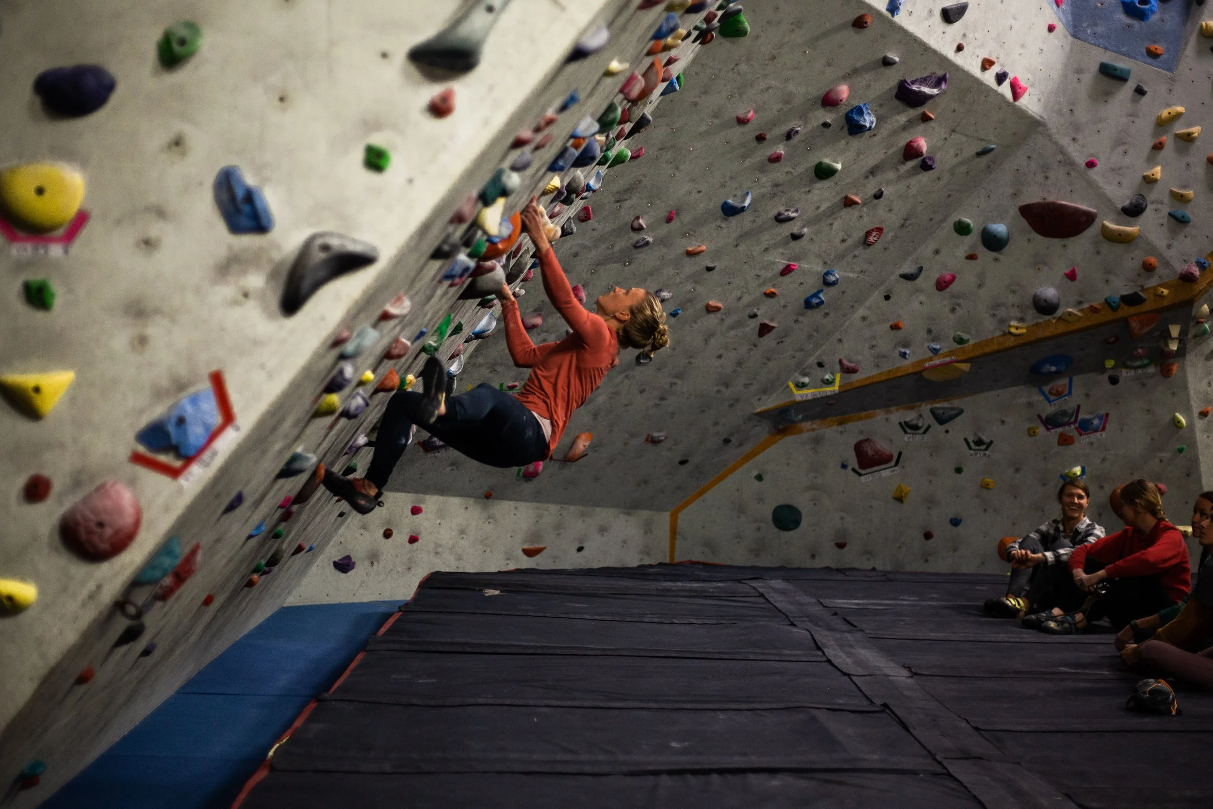 A woman climbing an indoor rock wall, with other people sitting and watching nearby.