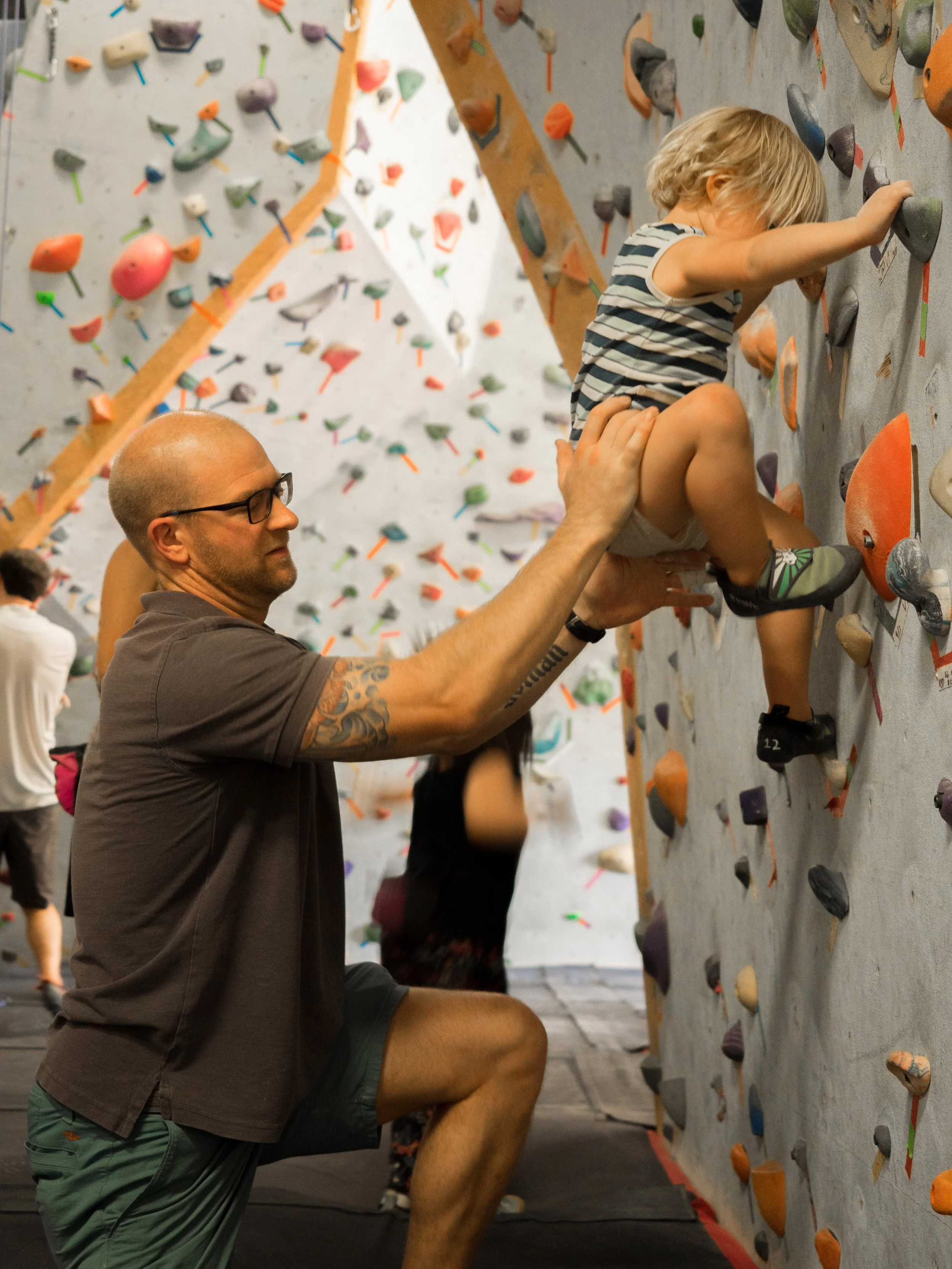 A man supporting a young girl climbing a rock wall at an indoor climbing gym.