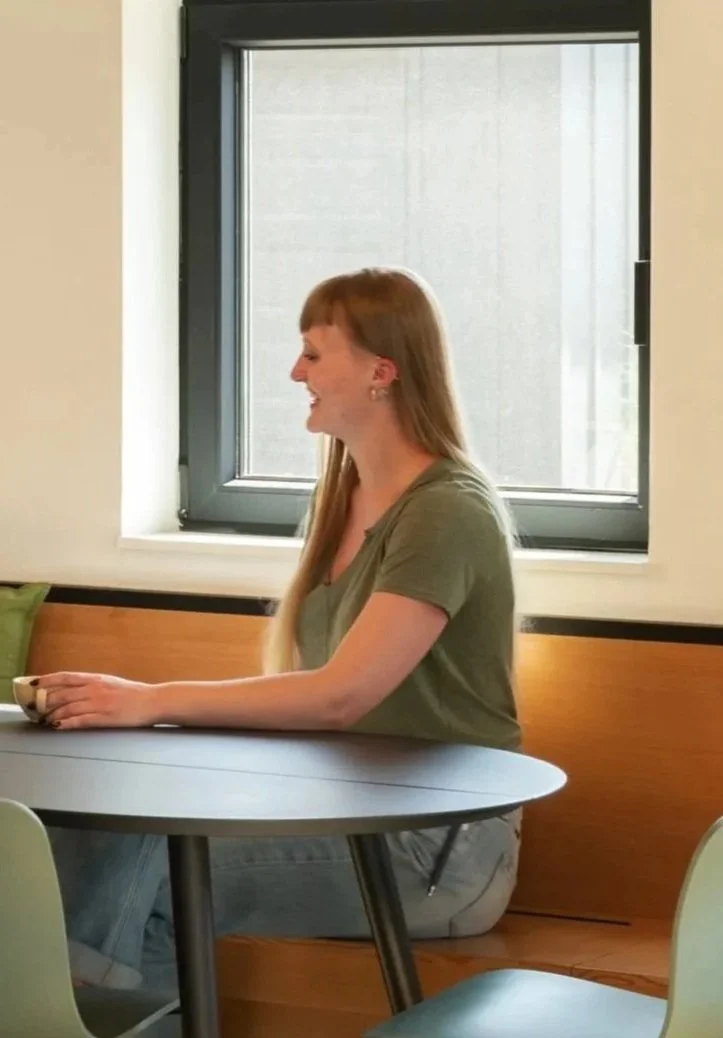 A woman with long reddish-brown hair, wearing a green t-shirt, sitting at a round table with a cup in her hand, smiling and looking to her left, in a room with a window in the background.