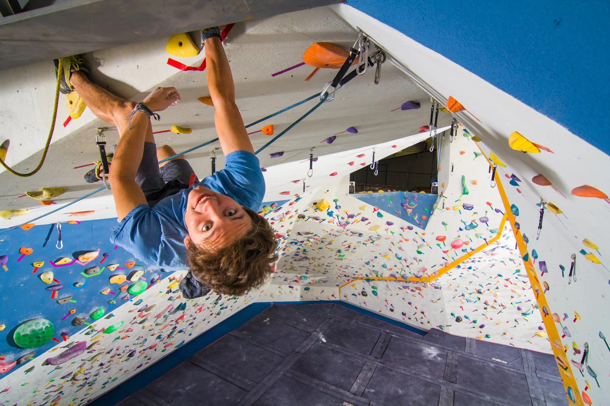 A person is climbing an indoor rock wall with colorful holds and safety gear, including harnesses and carabiners.