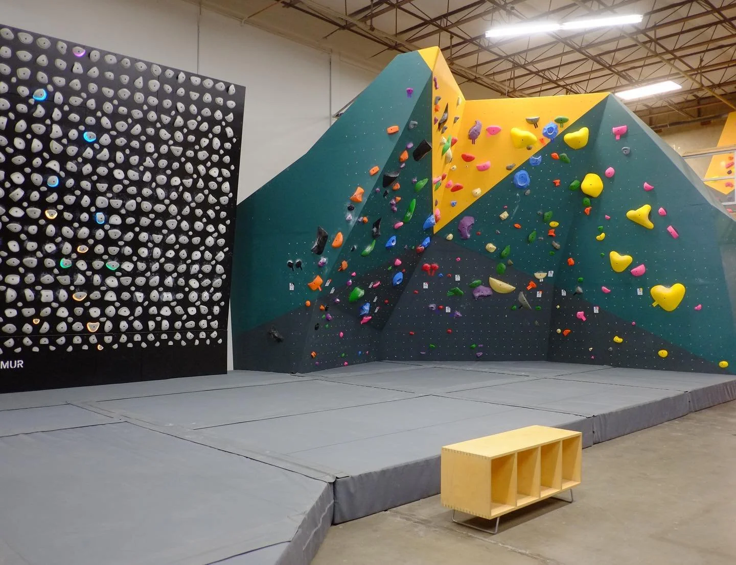 Indoor bouldering gym with black and yellow climbing walls filled with colorful holds, padded flooring, and a small wooden seating area.