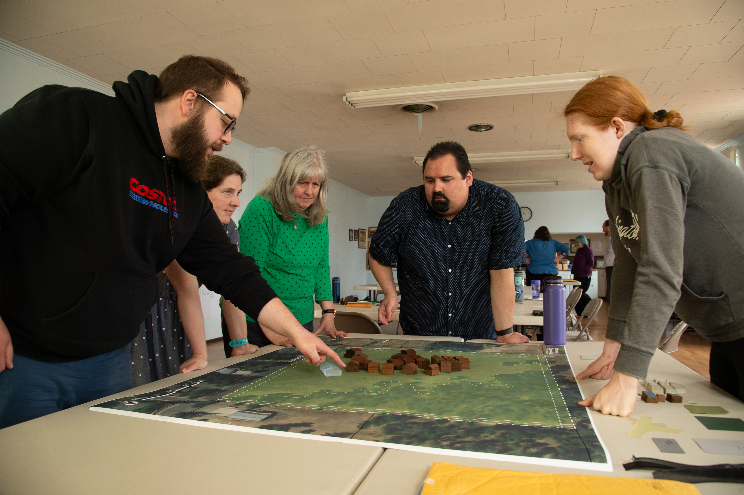 A group of five people gathered around a table, looking at a board game or map with wooden blocks on it, in a room with fluorescent lights and a clock on the wall.