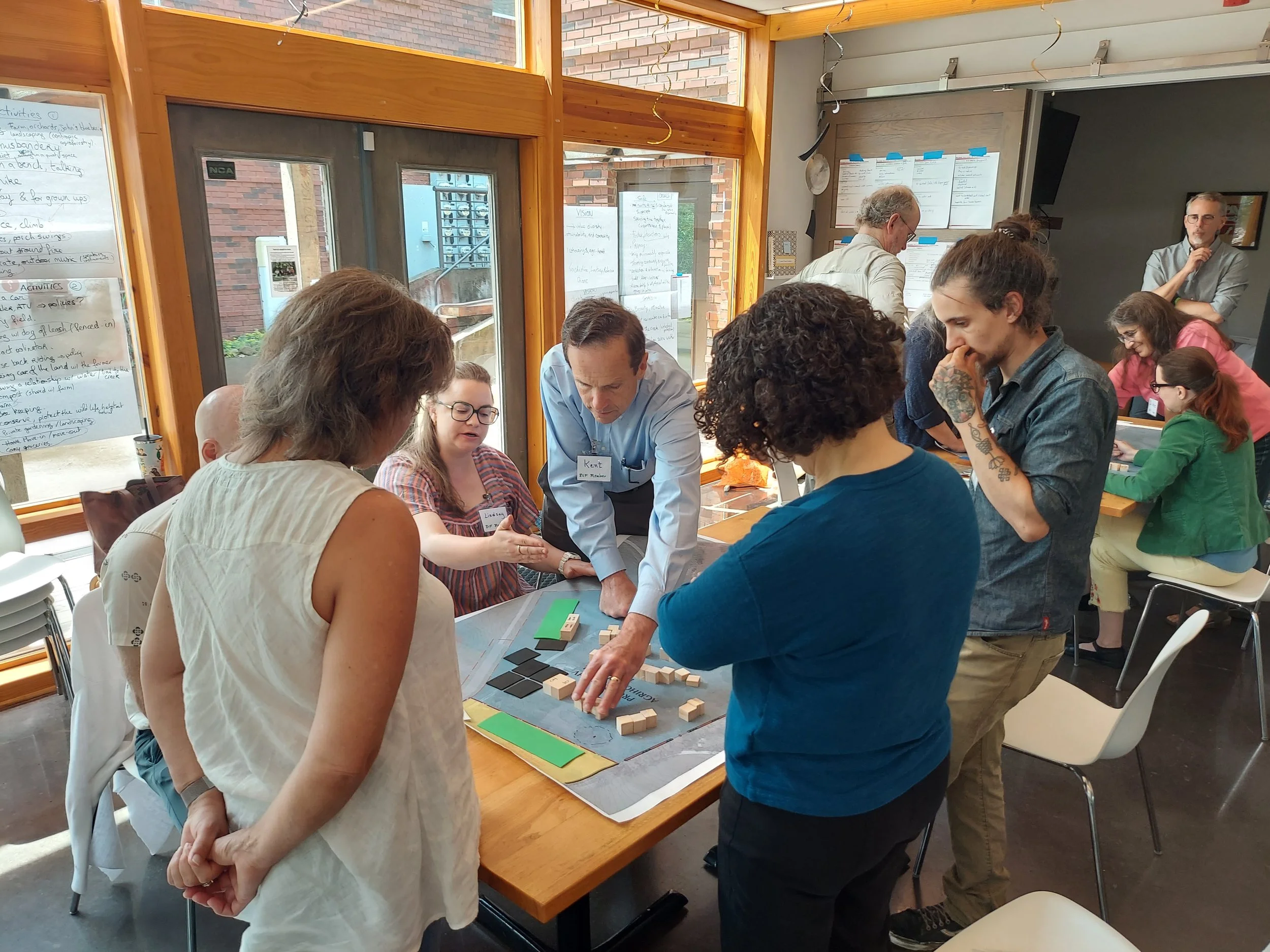 A group of people gathered around a table playing a tabletop game with wooden blocks and tiles on a board, in a room with large windows and notes on the wall.