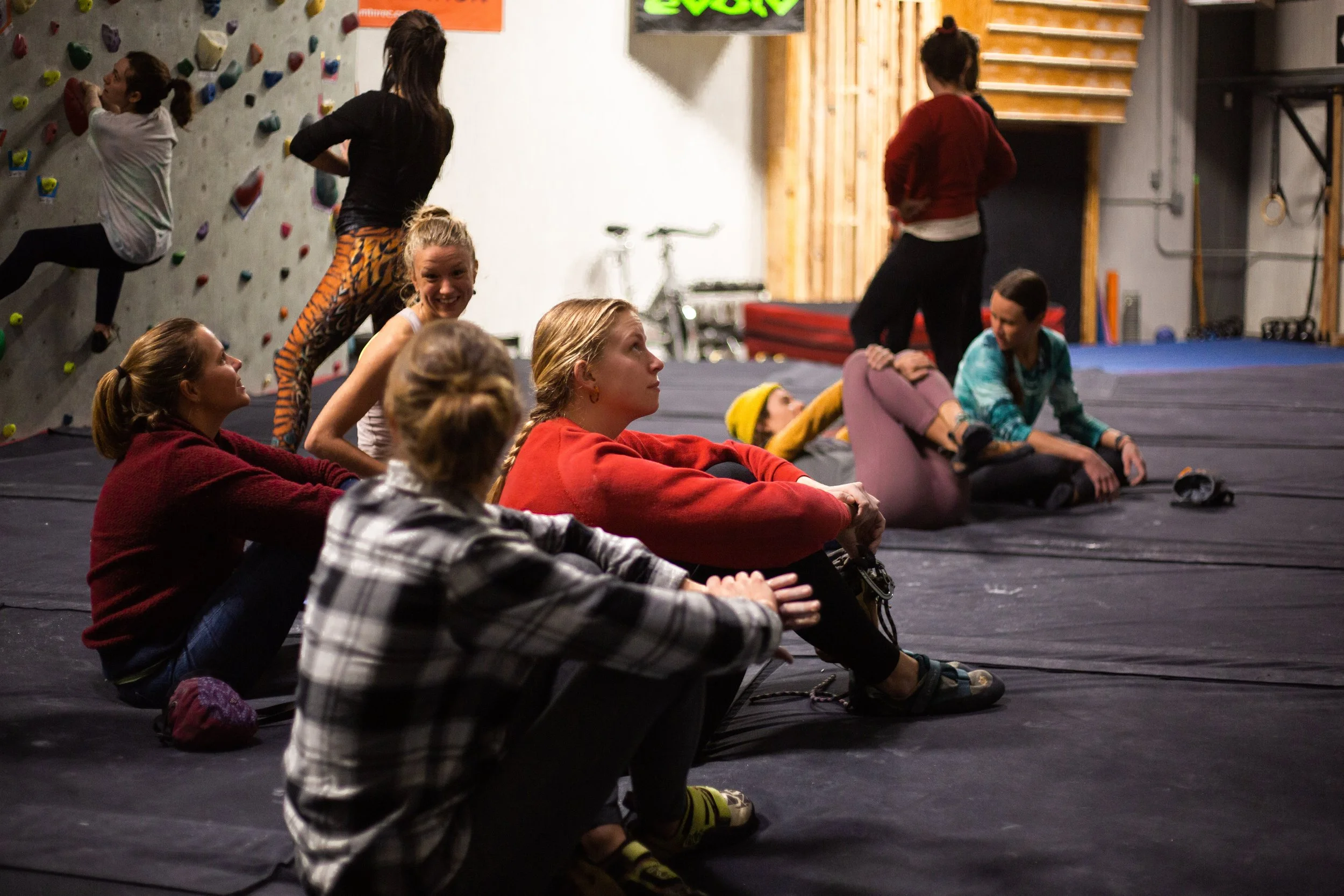 Group of young women relaxing on the floor of an indoor rock climbing gym, with some climbing on the wall in the background.