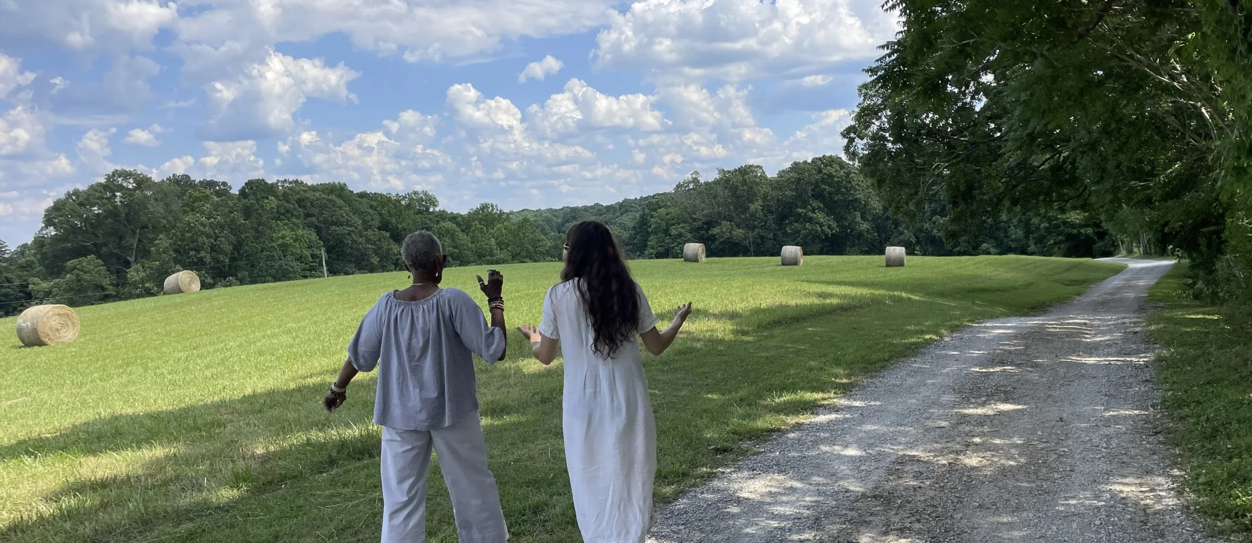 Two women walk along a dirt path in a grassy field with hay bales, surrounded by trees and a cloudy blue sky.