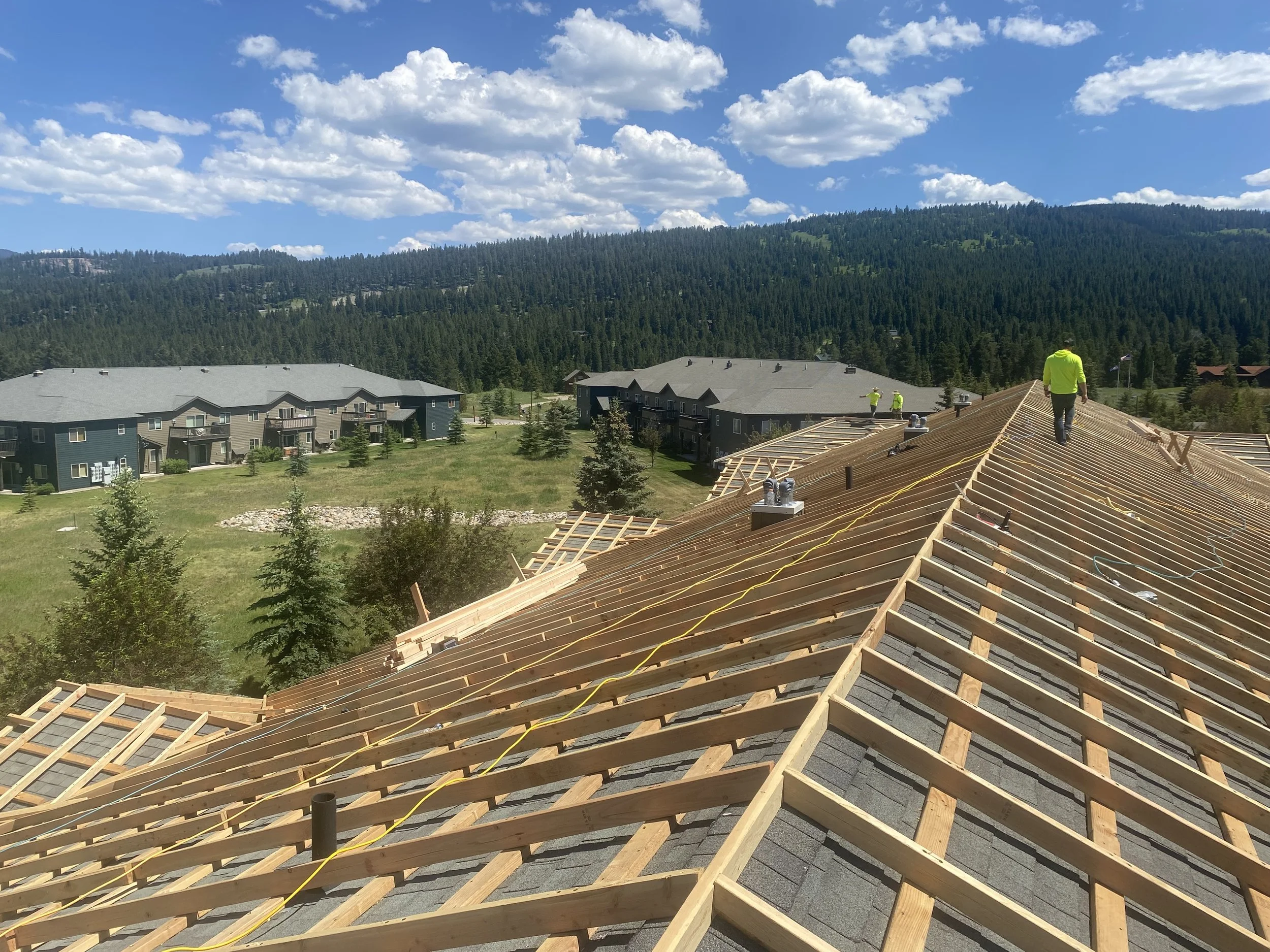 Construction workers on a roof under a clear blue sky with scattered clouds, in a residential area surrounded by green mountains and trees.
