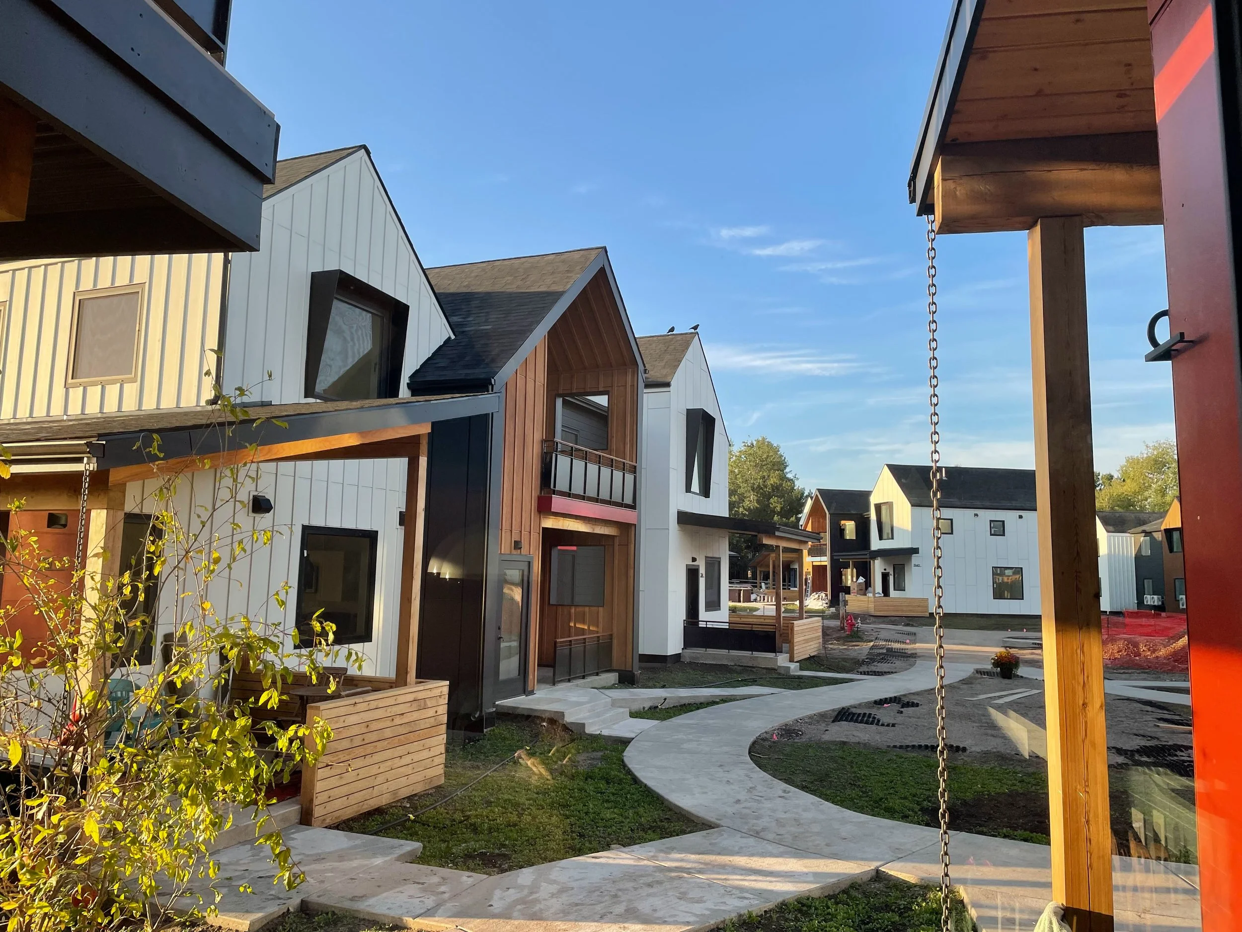 A neighborhood with modern, multi-story houses with different siding materials, including white, wood, and black. There are pathways, green lawns, and a swing set in the foreground on the right.