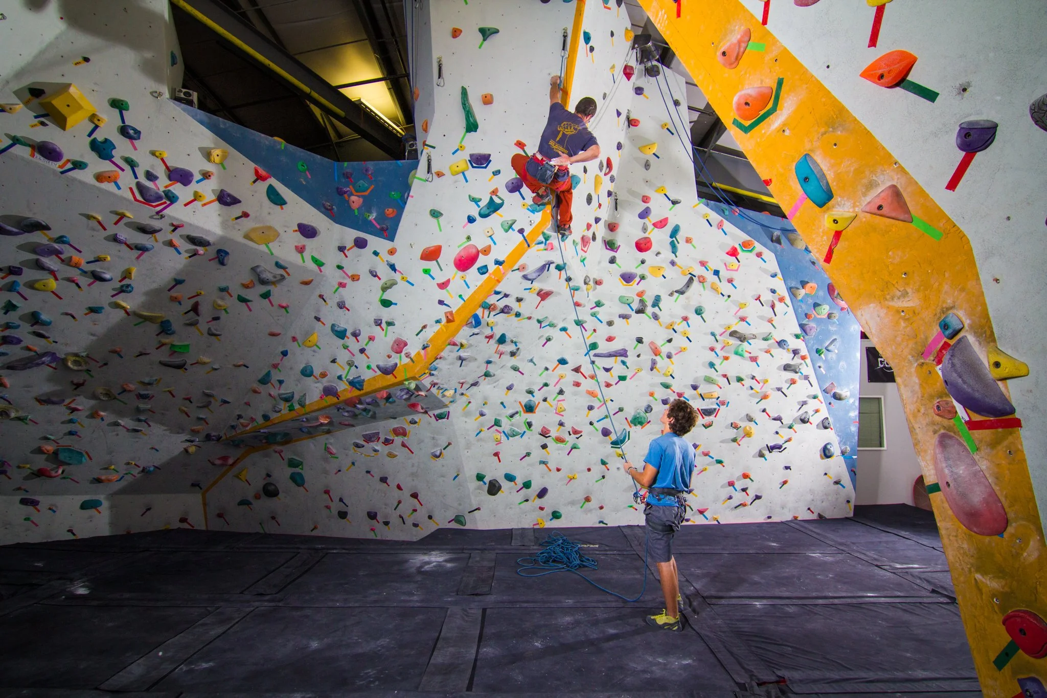 An indoor rock climbing gym with colorful climbing holds on the walls. A person is climbing while another is belaying from the ground.