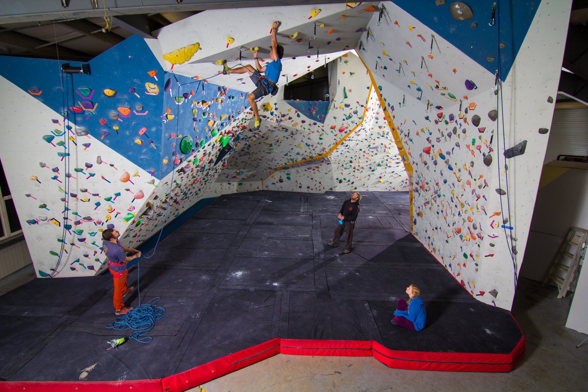 Indoor bouldering gym with colorful climbing holds on walls and overhangs, featuring a climber mid-ascent with safety harness, and spotters below.