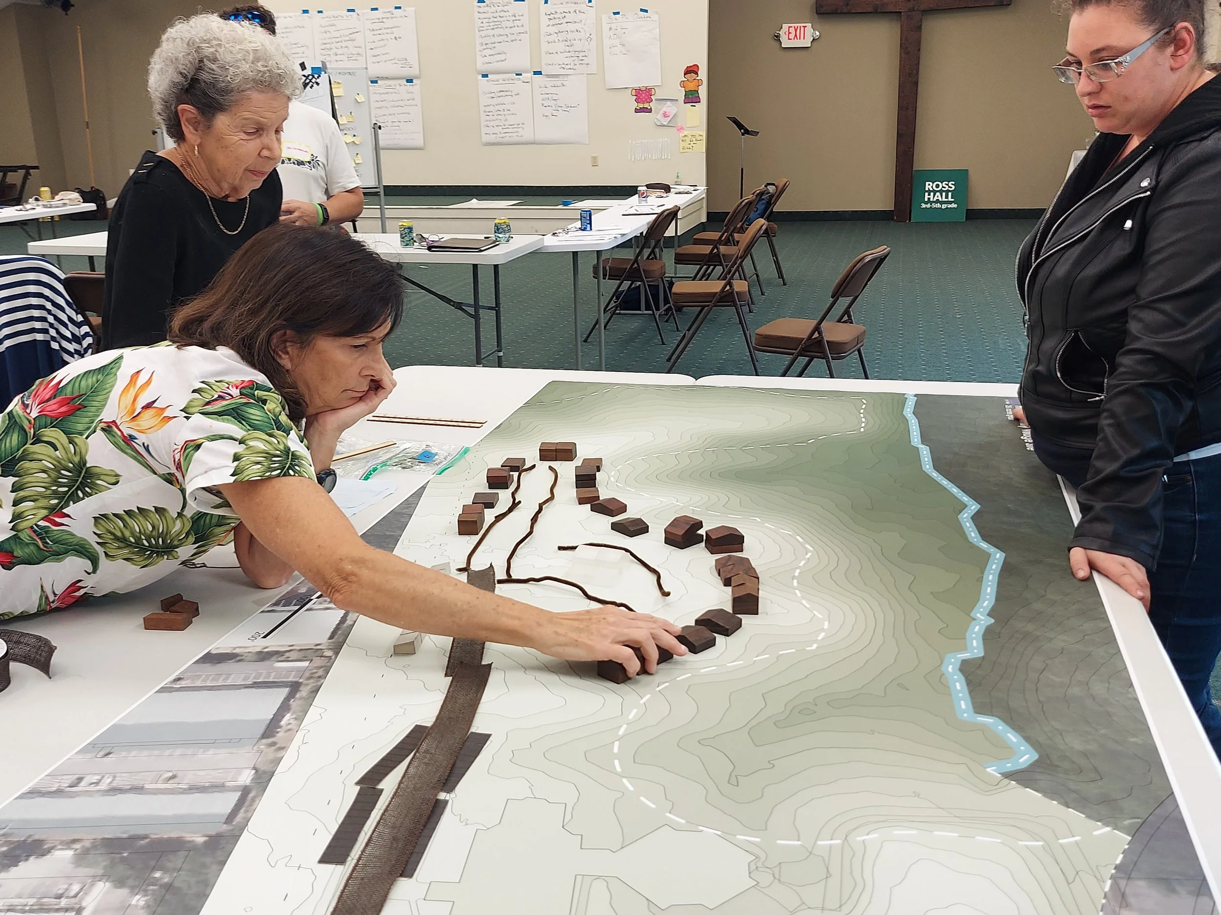 People gather around a large topographical map on a table, examining a layered landscape model with small building models and pathways, likely in a meeting or planning session.