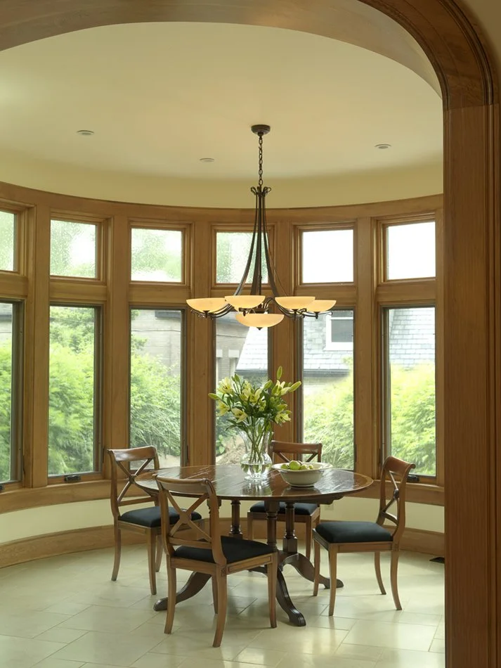 A dining area with a round wooden table, four chairs with black cushions, a glass vase with white lilies, and a chandelier hanging above. The space features large bay windows with a view of greenery outside.