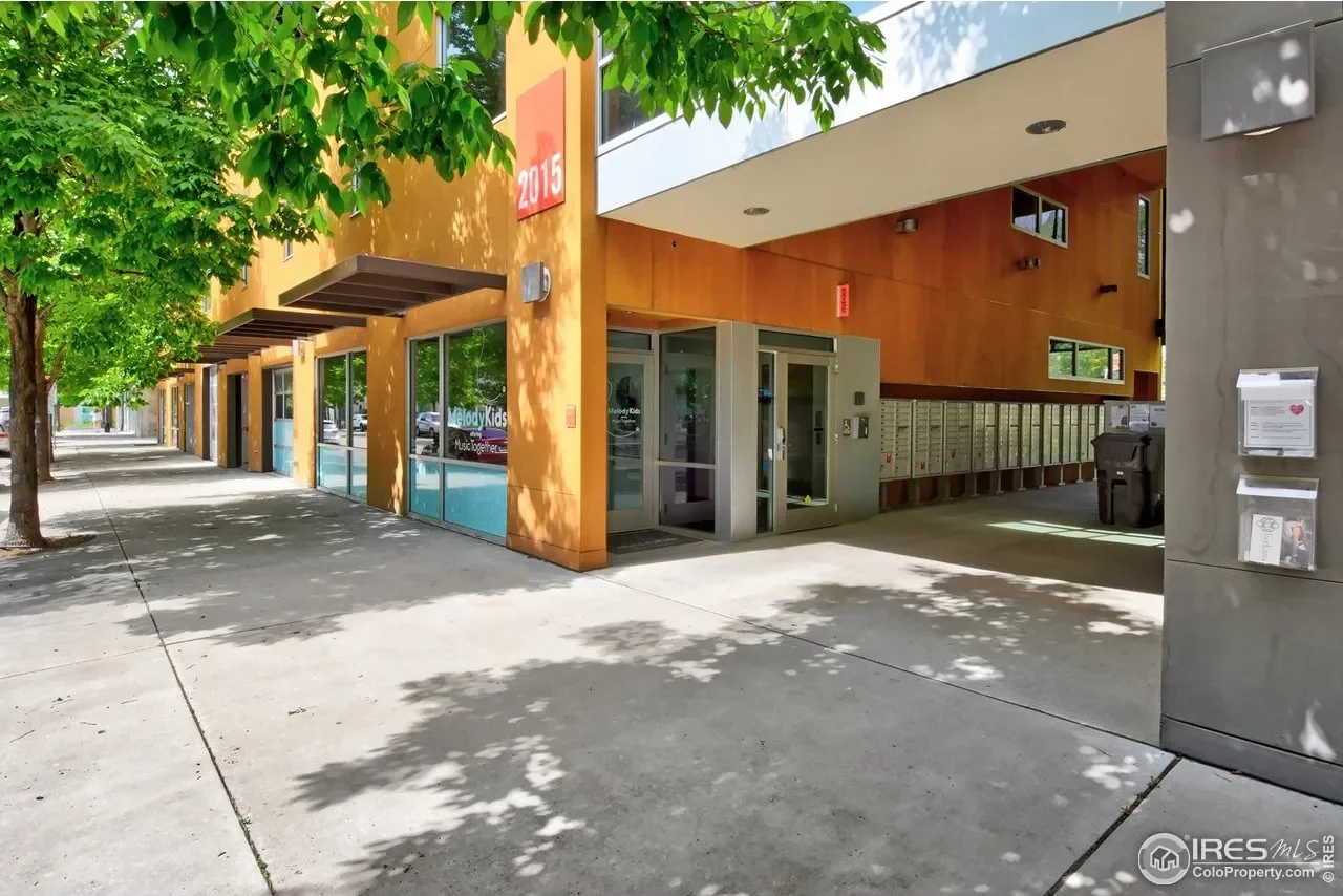Empty sidewalk in front of a modern commercial building with trees shading the area, including signage that says '2015', 'MelodyKids', and 'Music together'.