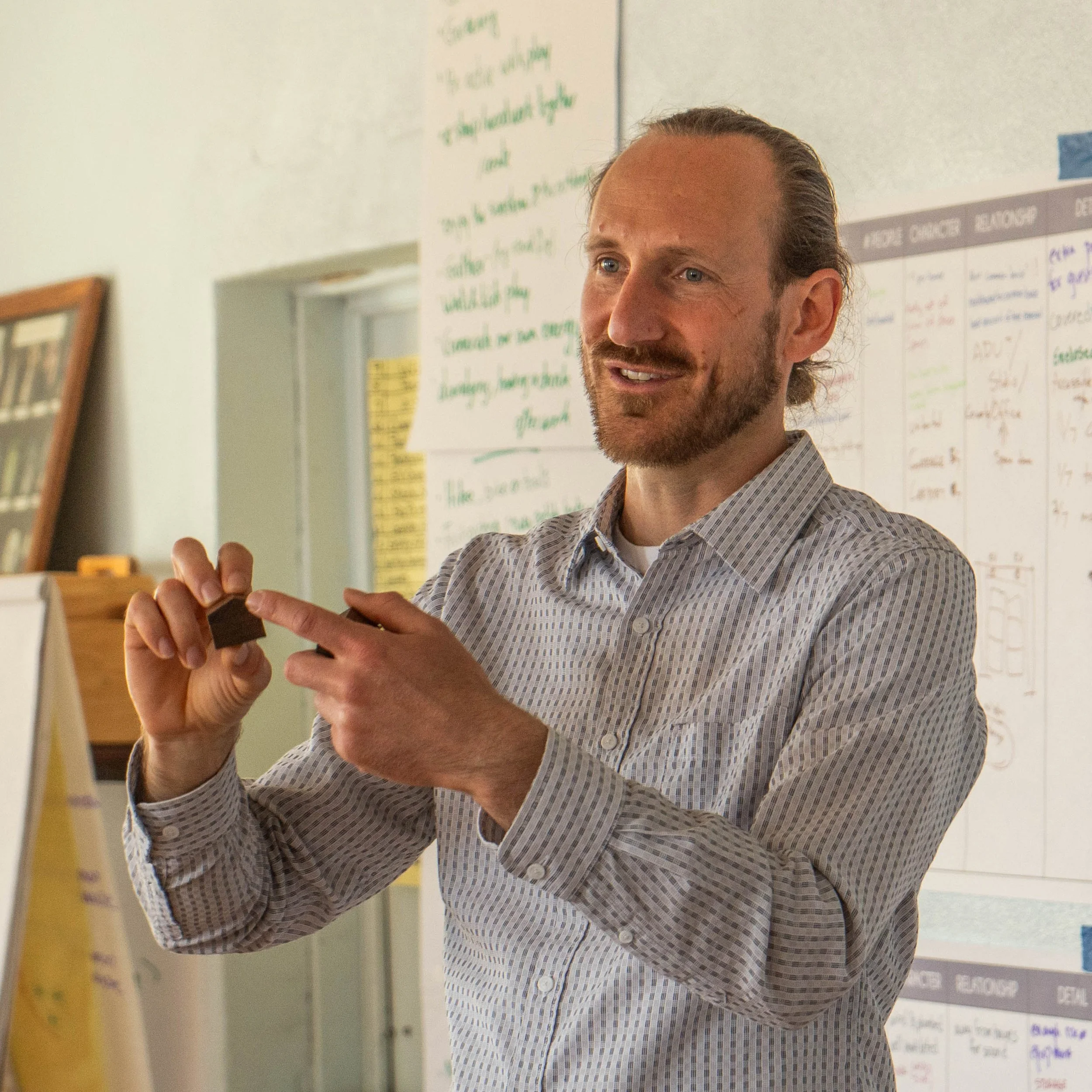 A man with a beard and tied-back hair is speaking and gesturing with his hand in front of a writable wall with notes and charts.