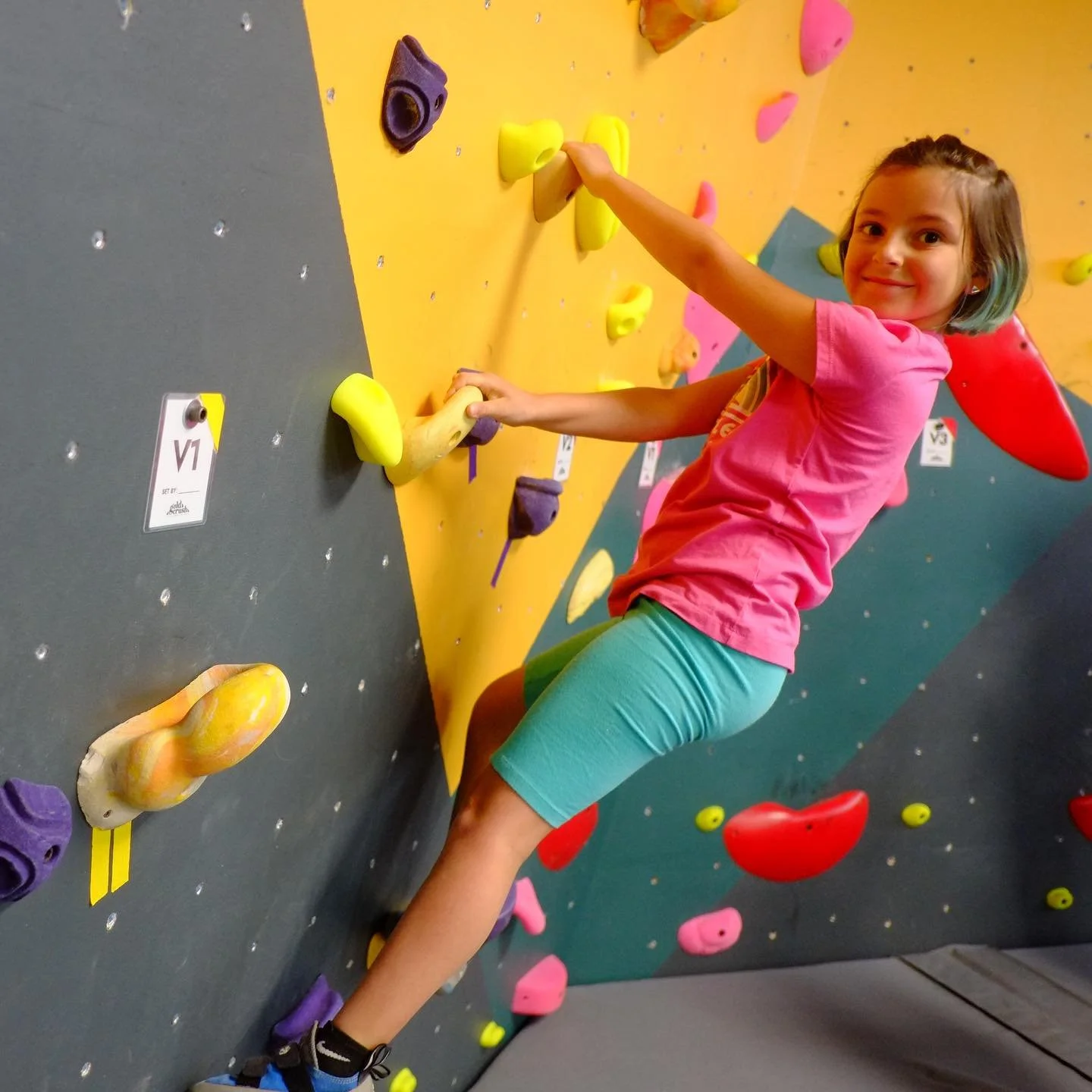 A young girl with short hair climbing a bouldering wall with colorful handholds, smiling at the camera.