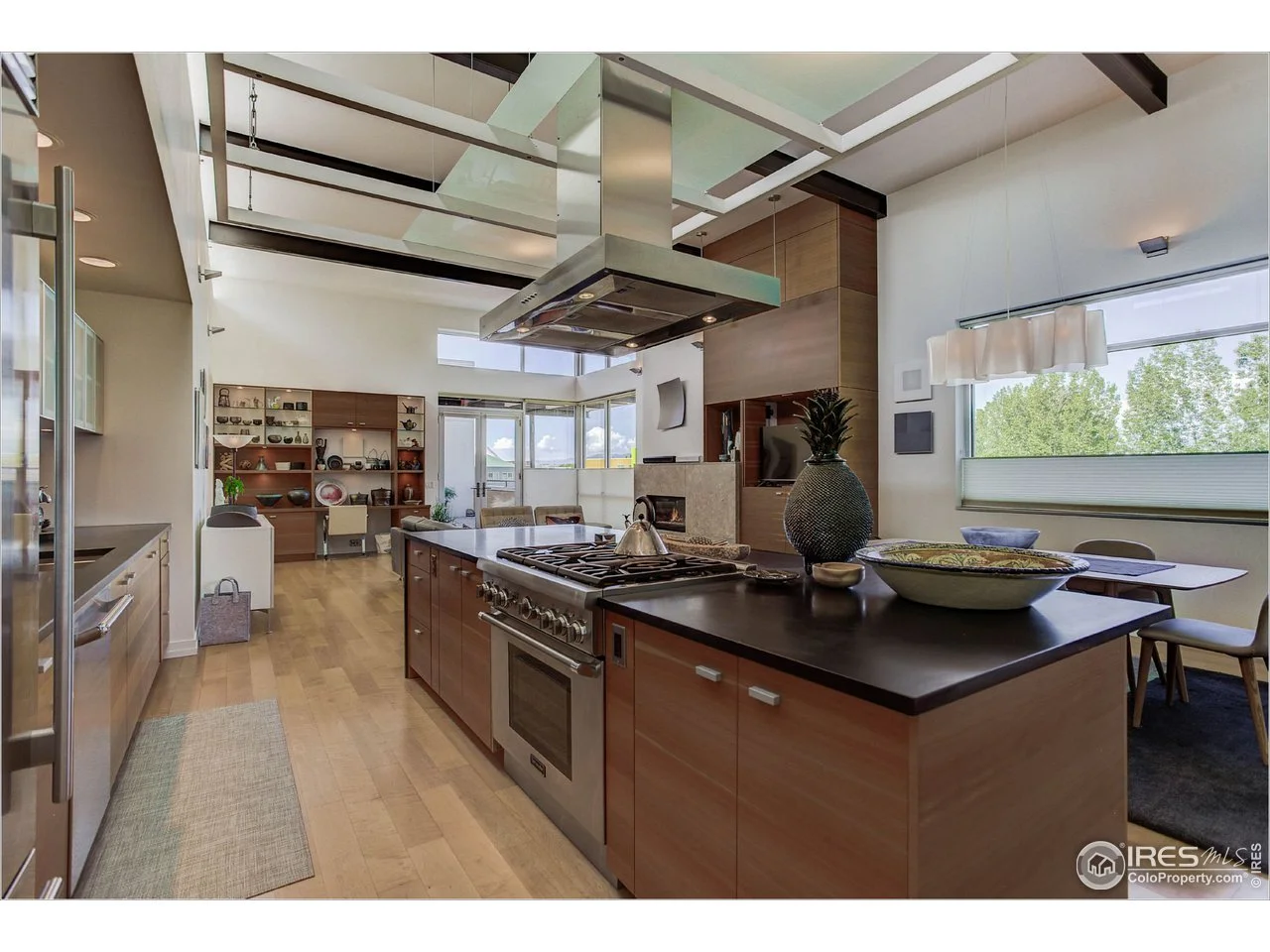 Open-concept kitchen with wooden cabinets, stainless steel appliances, a large island with a stovetop, and a dining area near windows showing greenery outside.