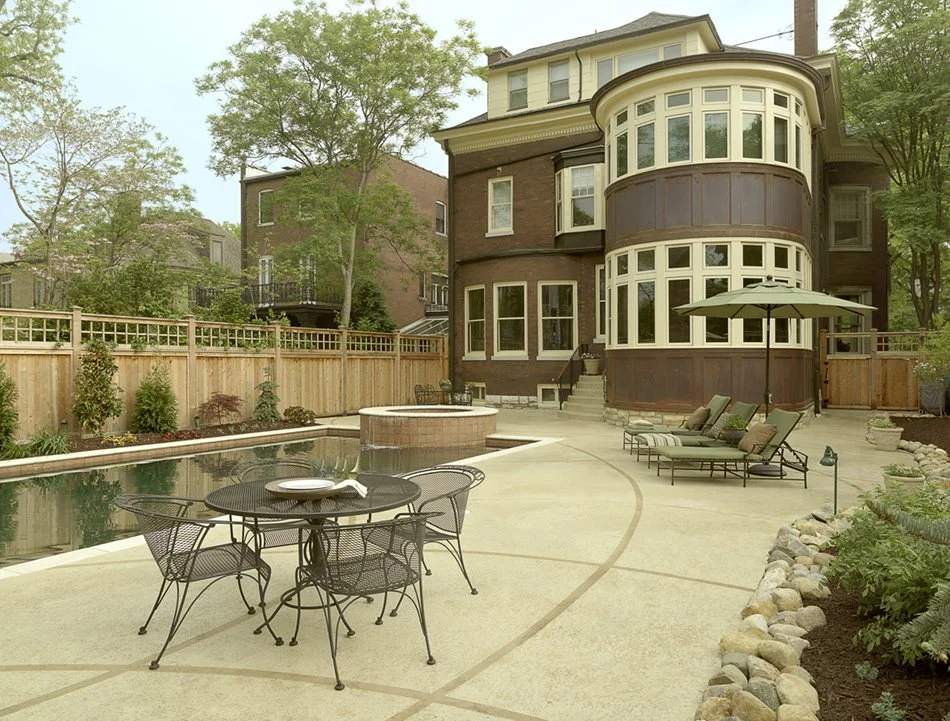 Backyard scene with a swimming pool, outdoor dining table, lounge chairs with cushions and umbrellas, and a multi-story brick house with rounded bay windows, surrounded by a wooden fence and greenery.