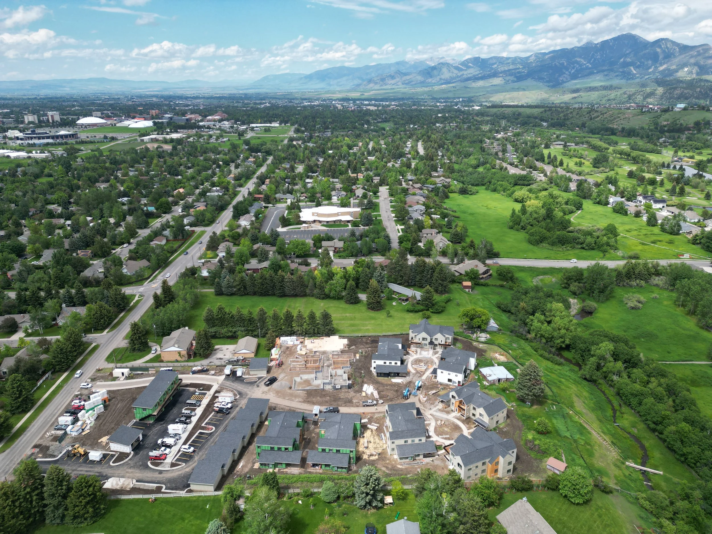 An aerial view of a suburban neighborhood with houses, trees, and construction sites, with mountains in the background under a partly cloudy sky.