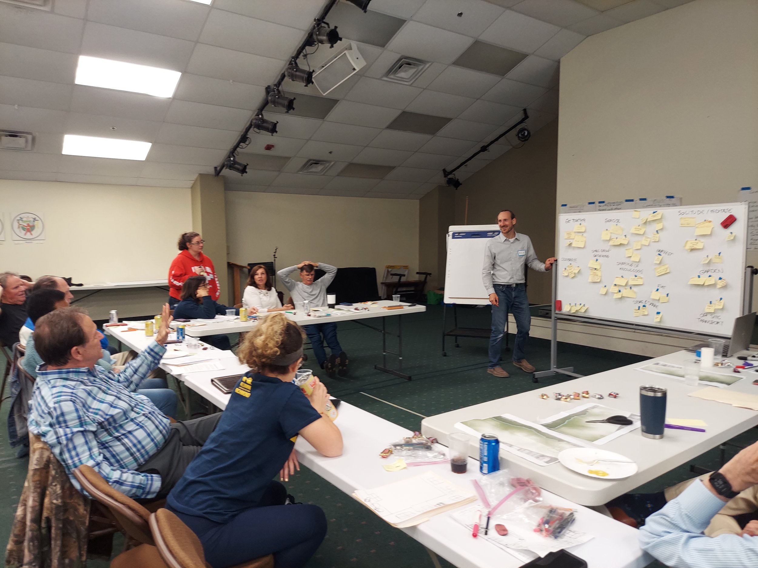 A group of people sitting around tables in a conference room, with a man standing in front near a whiteboard filled with sticky notes, during a workshop or brainstorming session.