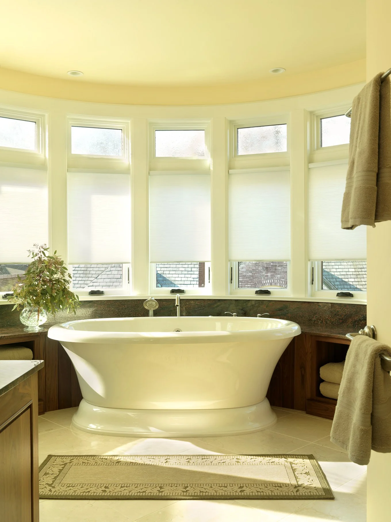 A bathroom with a freestanding bathtub in front of a large bay window with multiple small panes, white blinds, and a view of rooftops outside. There is a plant in a glass vase on a dark countertop to the left of the tub, and towels on a towel rack an