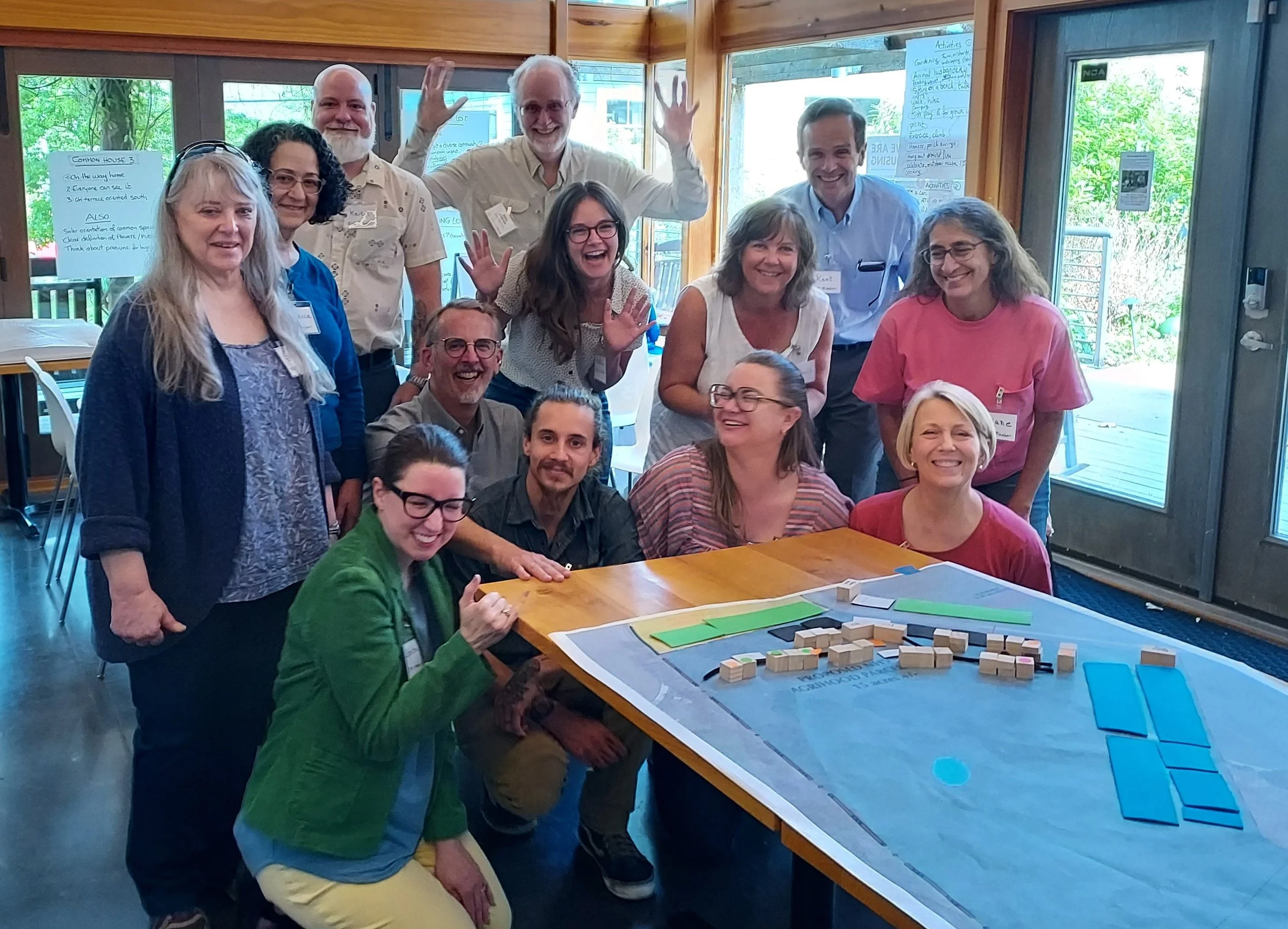 A group of smiling people, some standing and some sitting, gathered around a table with a large map and small wooden blocks, in a well-lit room with wood-paneled walls and large windows showing greenery outside.