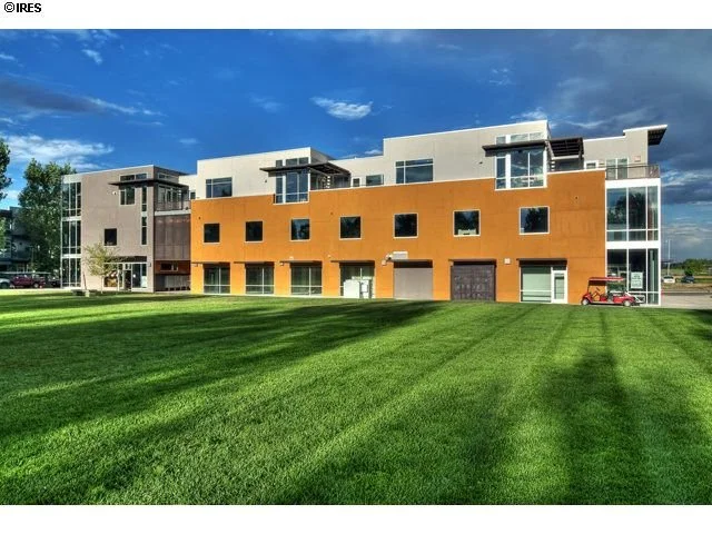 Modern apartment building with a combination of white, gray, and orange facades, large windows, and a well-manicured green lawn in front.