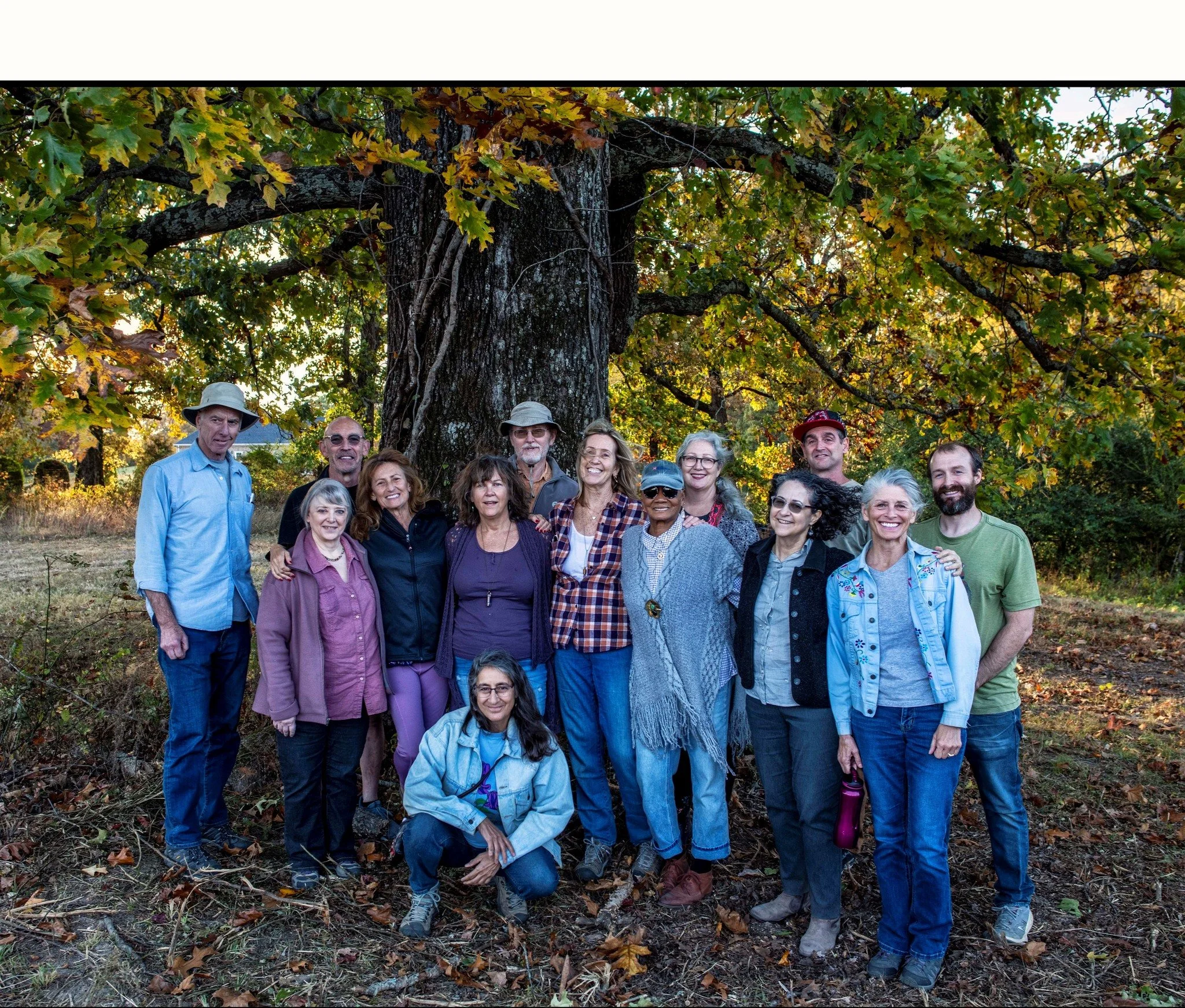 A group of diverse people gathered outdoors under a large leafy tree during autumn, smiling and posing for a photo.