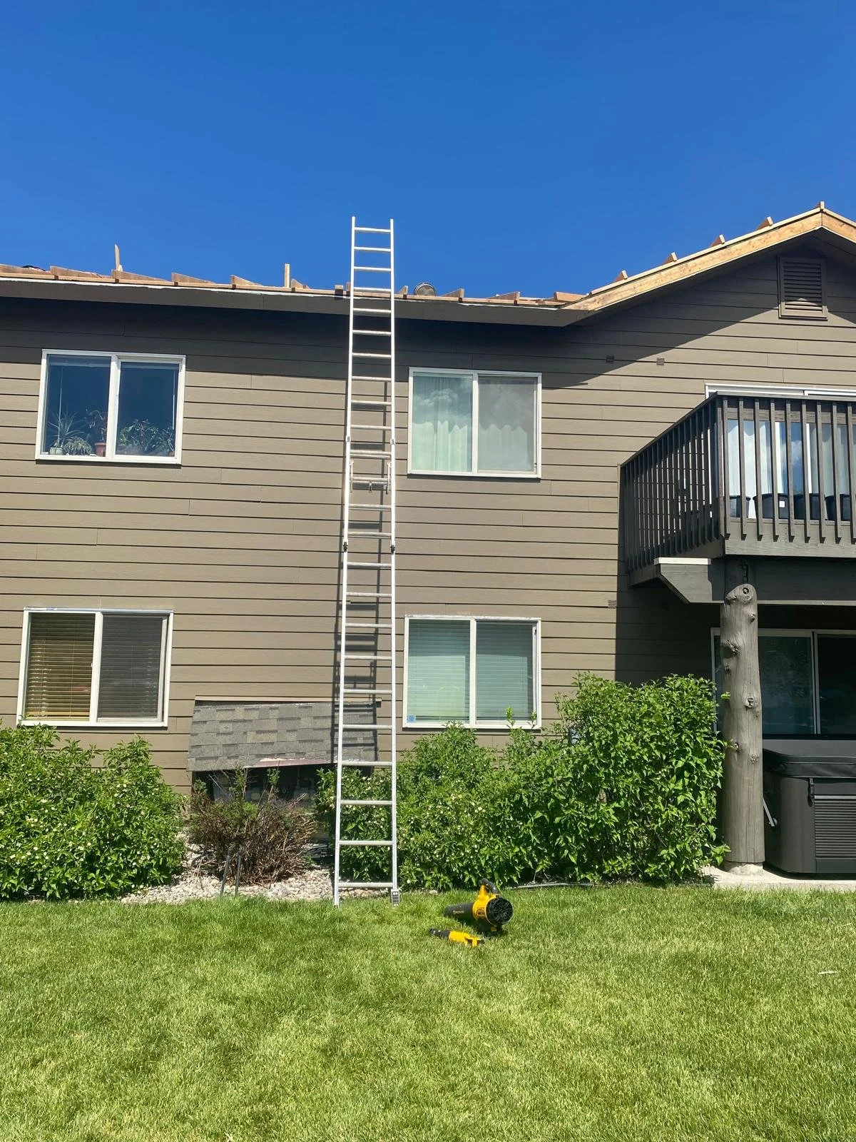 Side of a residential building with a metal ladder leaning against the roof, four windows, a small balcony, bushes, a lawn, and landscaping tools on the grass.