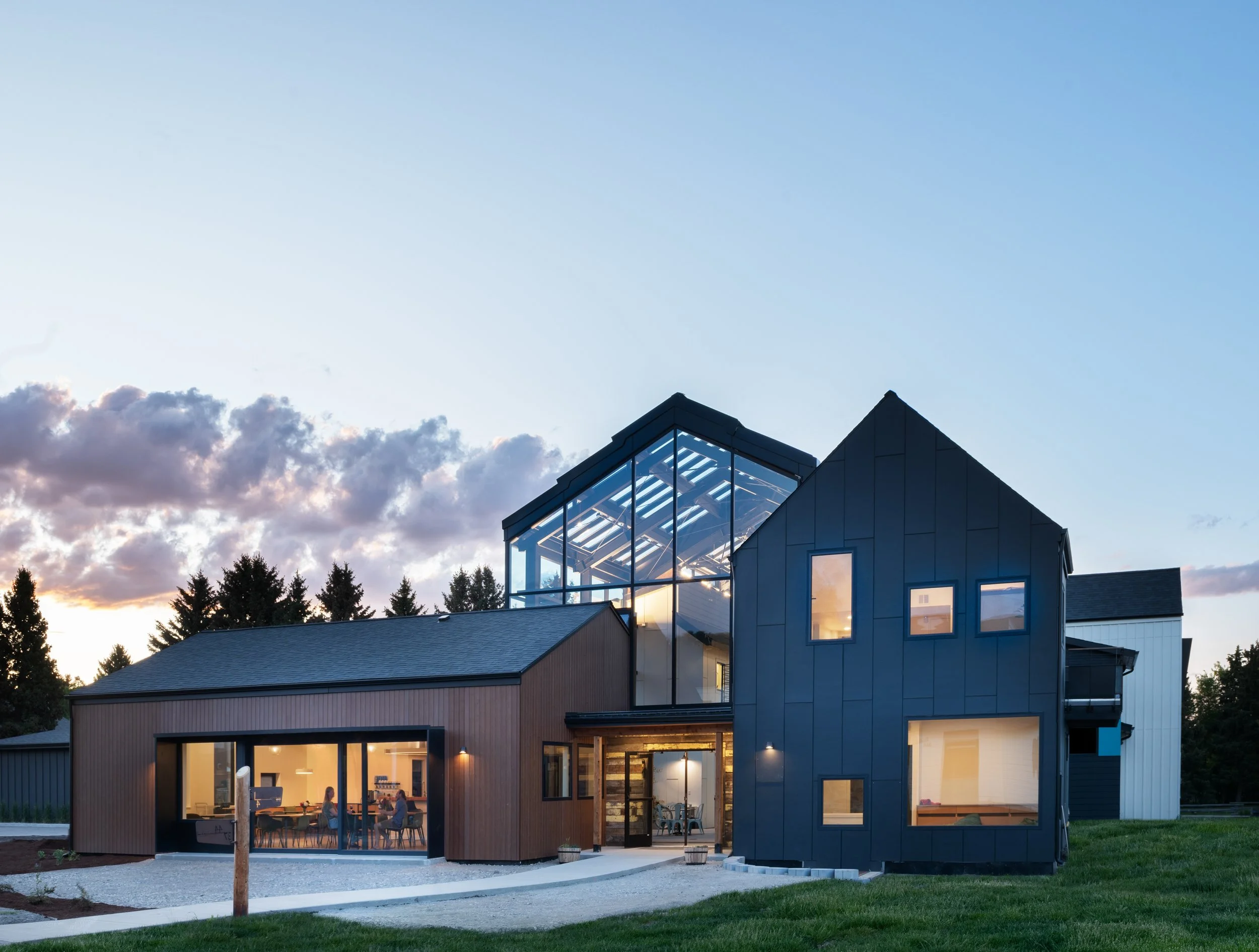 Modern house with large glass windows and a rooftop greenhouse, illuminated from inside, with a blue sky and clouds in the background.
