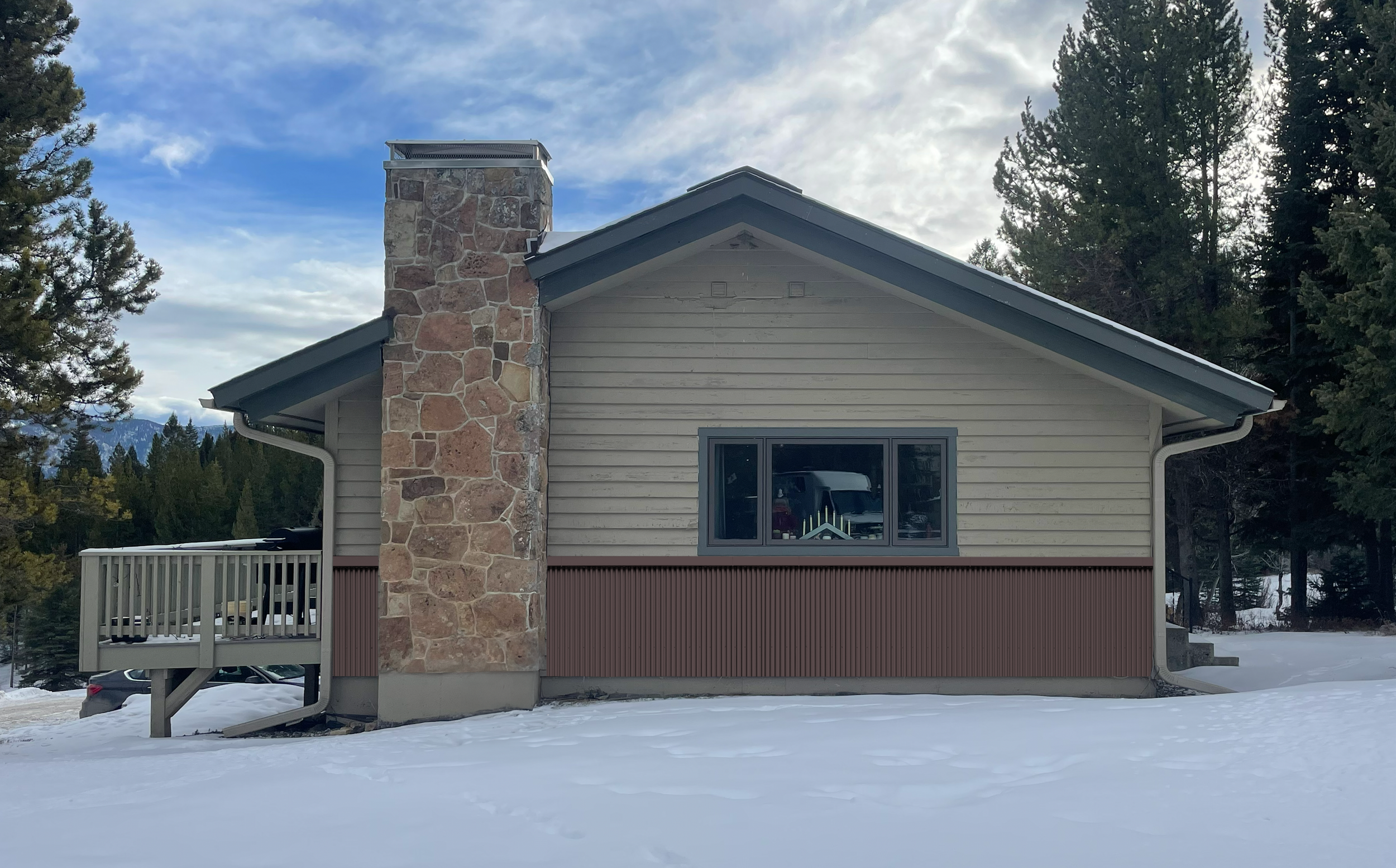 View of the back of a house with beige siding, a stone chimney, a window reflecting the outdoors, a white railing, and snow-covered ground surrounded by evergreen trees under a partly cloudy sky.
