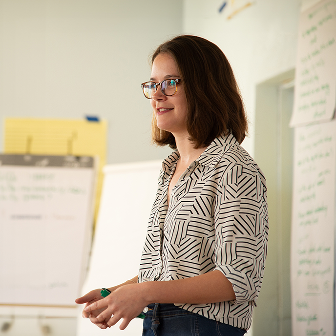 A woman with shoulder-length brown hair, glasses, and a patterned blouse standing in a bright room with white walls and charts.