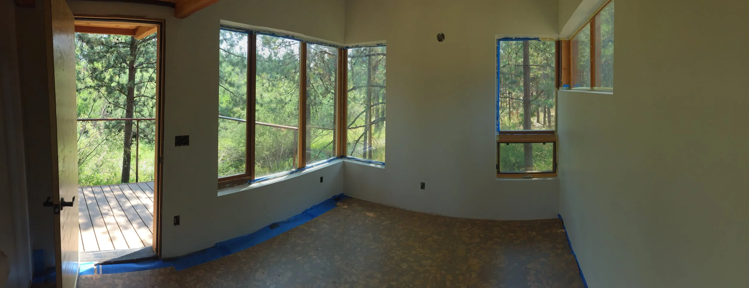 Room under construction with multiple large windows showing a view of trees outside, blue painter's tape along window edges and base of walls, and a partially finished wooden French door leading to a deck.