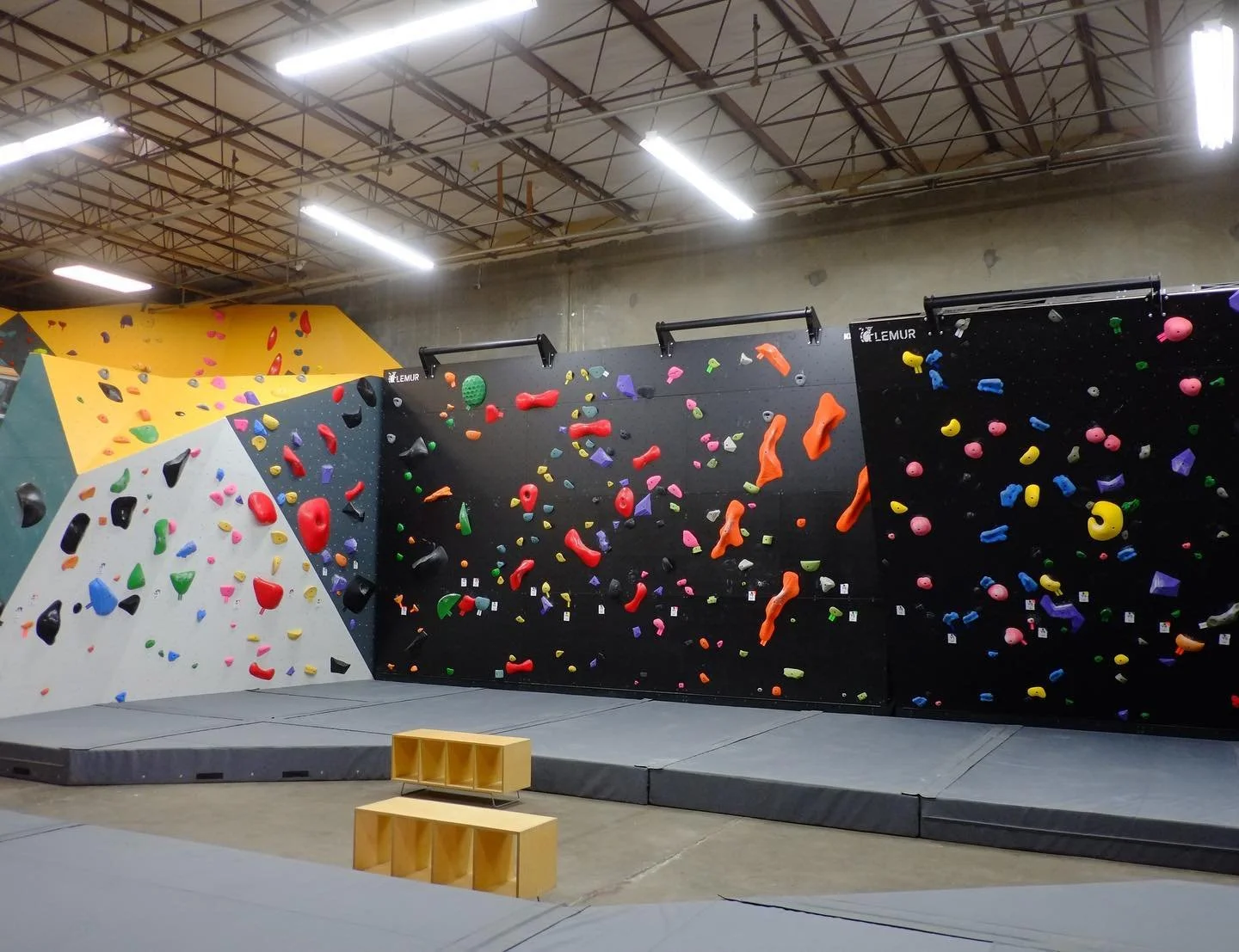 Indoor bouldering gym with black and white walls covered in colorful climbing holds, padded floor, and wooden boxes in the foreground.