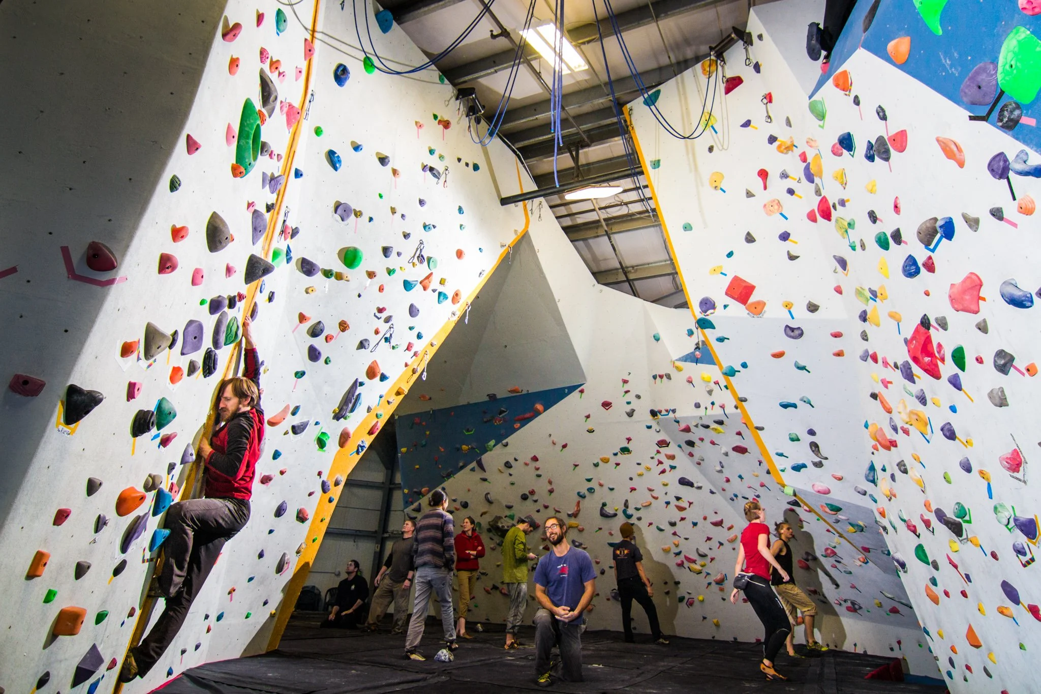 Indoor rock climbing gym with colorful holds and multiple climbers, some ascending the wall and others watching.