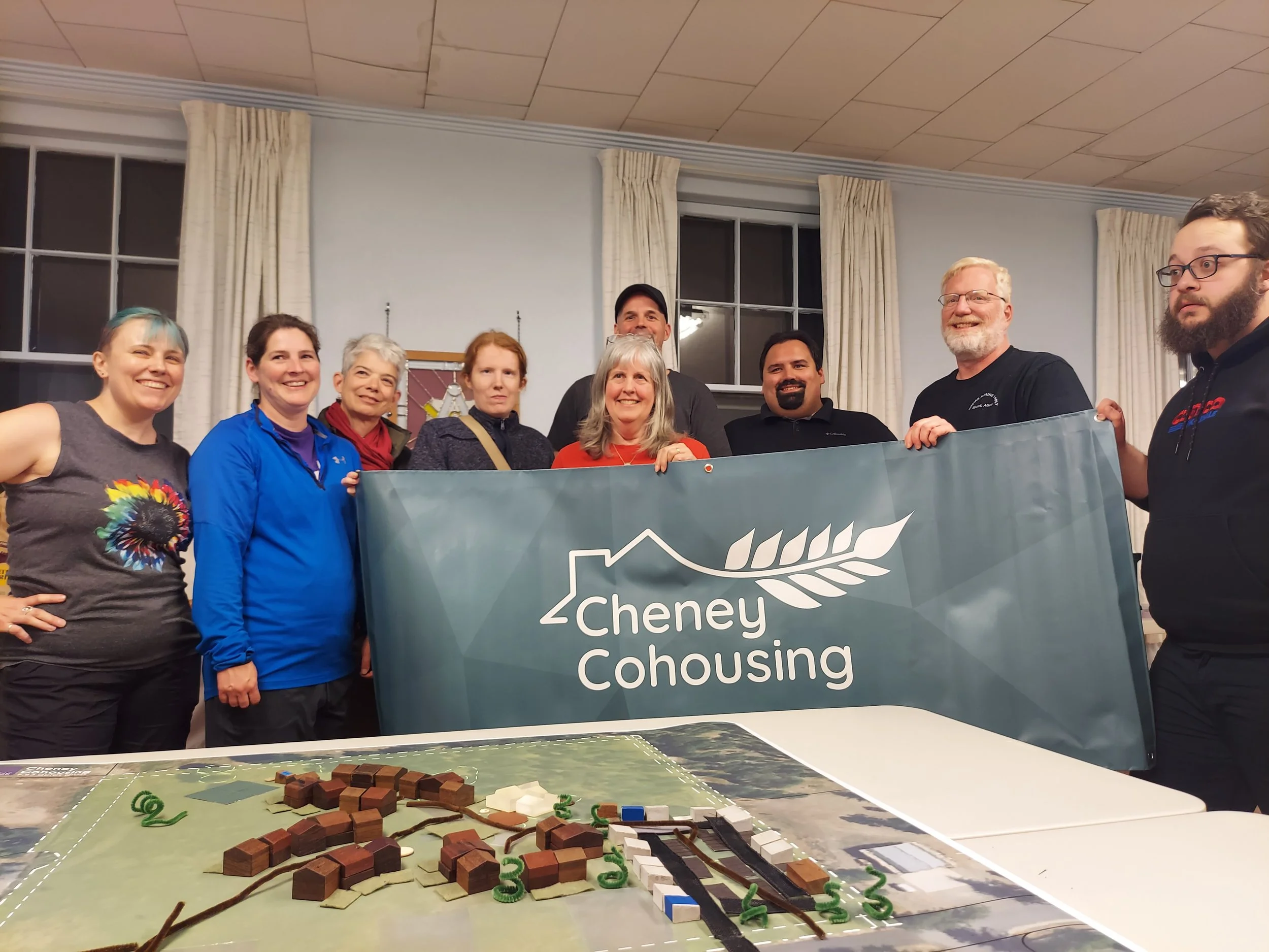 Group of diverse people holding a banner that says Cheney Cohousing, standing behind a table with a detailed scale model of a housing development, in a room with large windows and curtains.