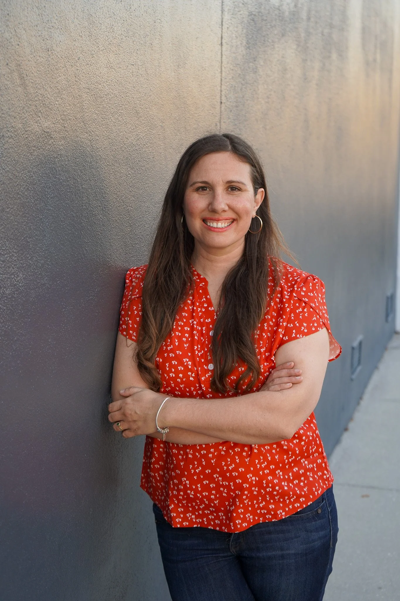 Author Katie Parsons leans against a wall. She is wearing an orange short-sleeved shirt.