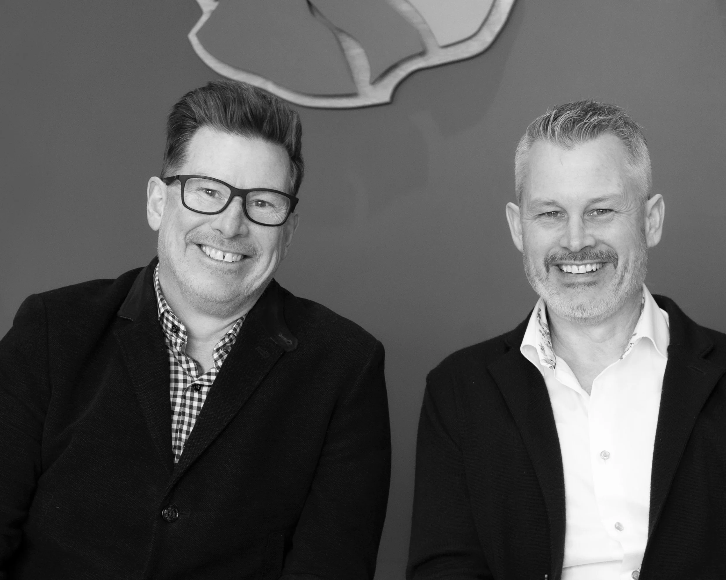 Black and white photo of two smiling men in business attire sitting side by side, on a plain wall background.
