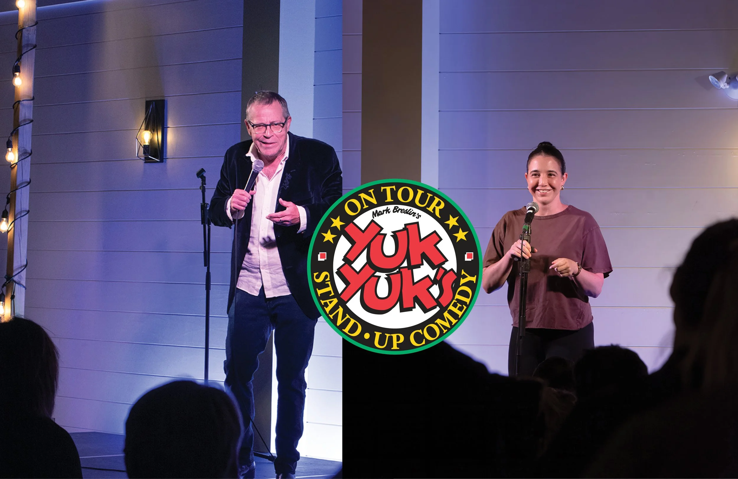 A comedy show stage with two performers, a man and a woman, standing in front of an audience. The man is wearing glasses, a black velvet blazer, and is speaking into a microphone. The woman is smiling, wearing a brown top, and also holding a microphone. There is a circular logo overlay in the center of the image that reads "Yuk Yuk" with additional text indicating it is a stand-up comedy tour featuring Mark Breslin.