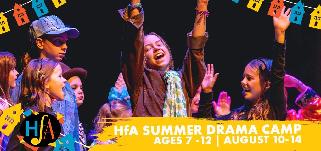 Children participating in a summer drama camp, wearing costumes and colorful accessories, smiling and celebrating on stage with decorations of hanging paper houses in the background.