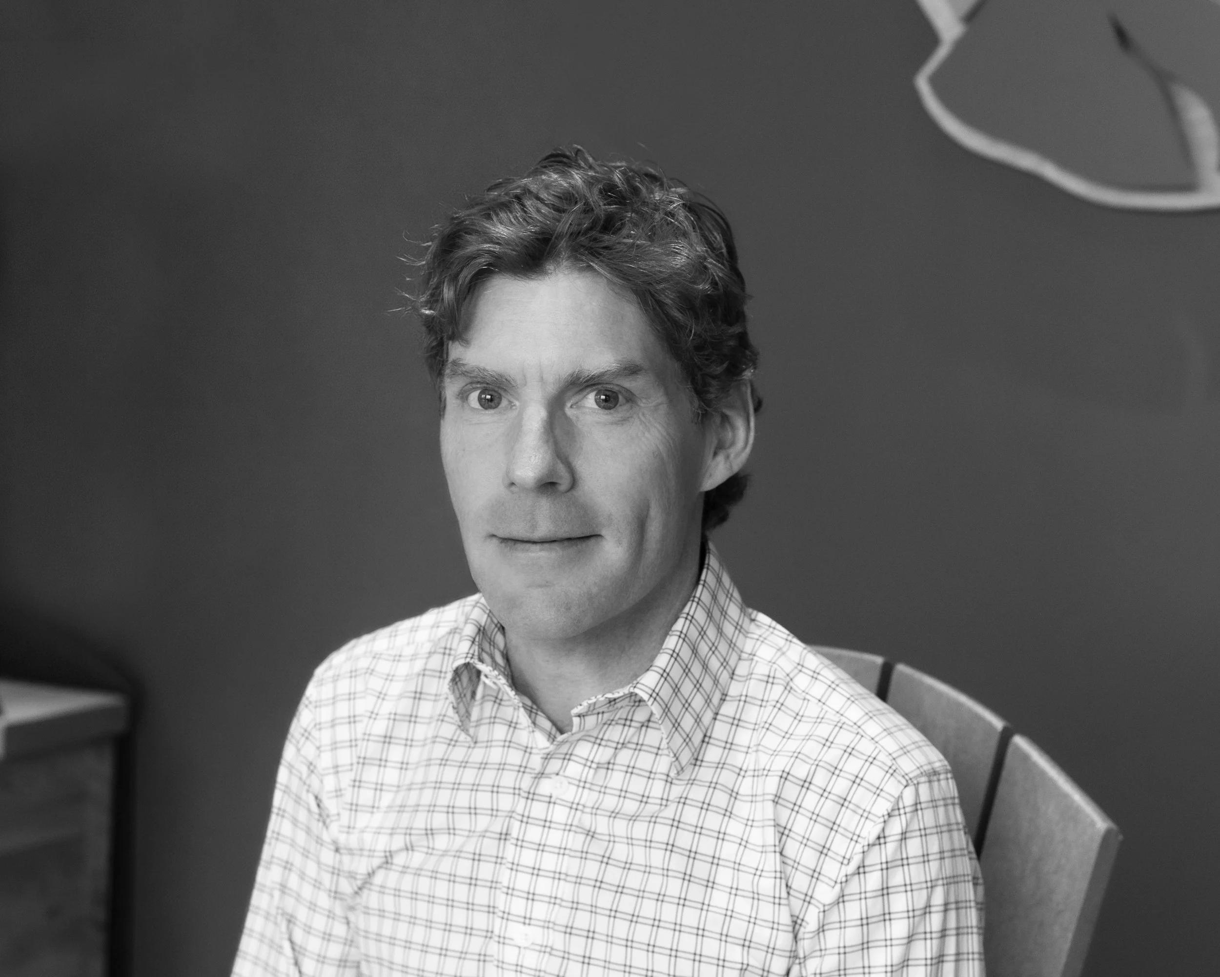 Black and white portrait of a middle-aged man with wavy hair, wearing a checkered button-up shirt, sitting on a chair with a slight smile, against a plain background.