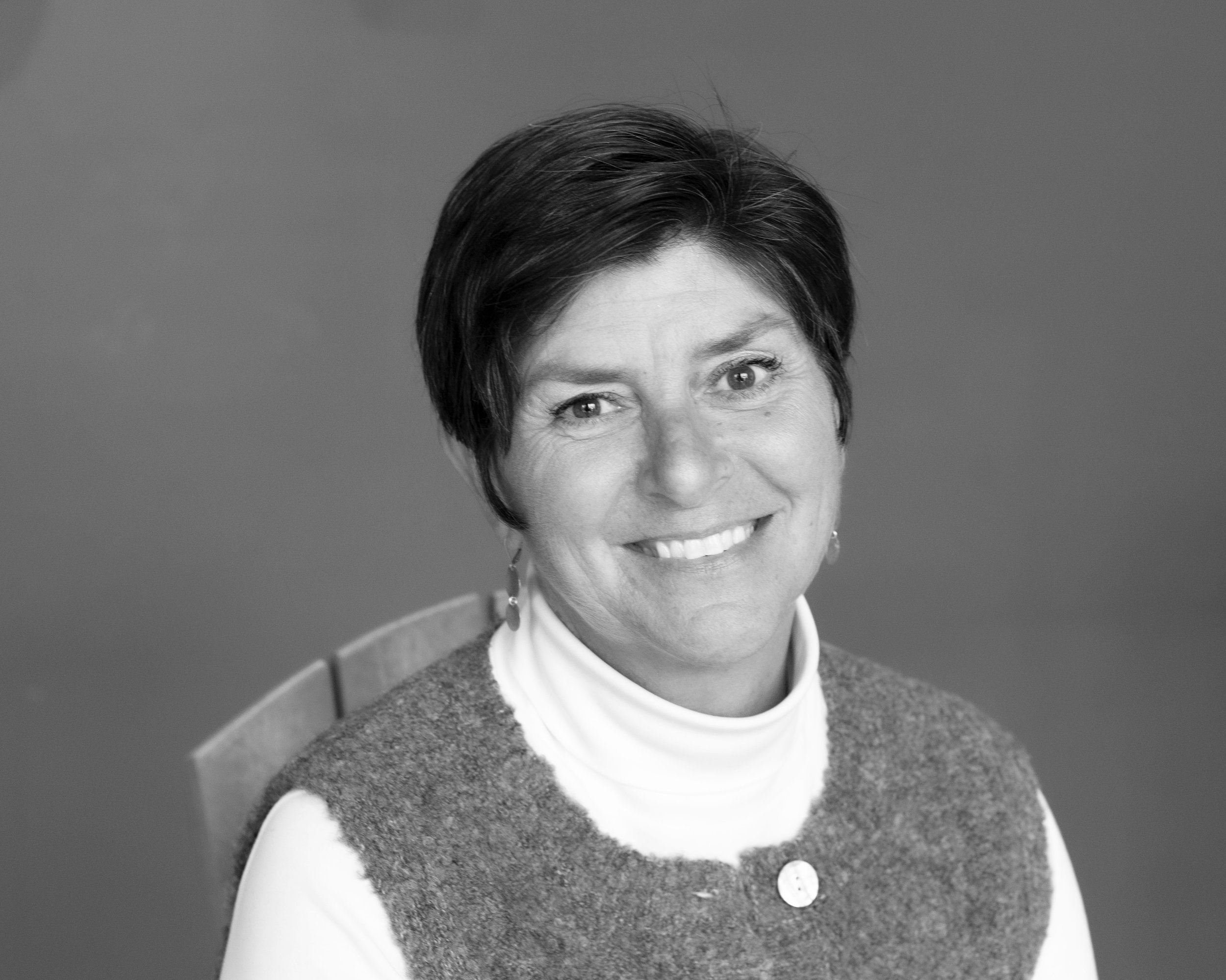 Black and white portrait of a woman with short dark hair, smiling, wearing earrings, a white turtleneck, and a textured vest, sitting on a wooden chair against a plain background.
