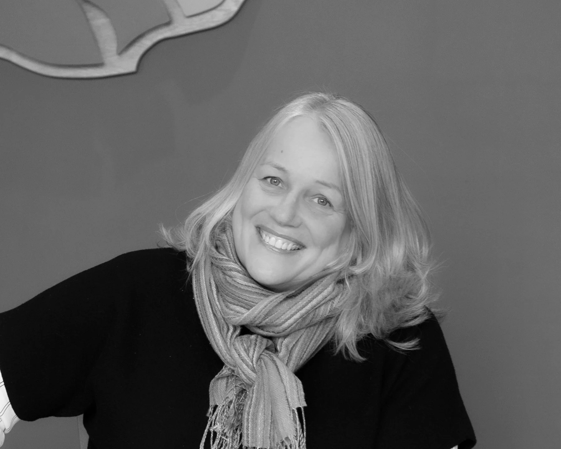 A woman with shoulder-length wavy hair smiling, wearing a black top and a striped scarf, sitting indoors against a plain wall with a decorative object hanging on the upper left corner.