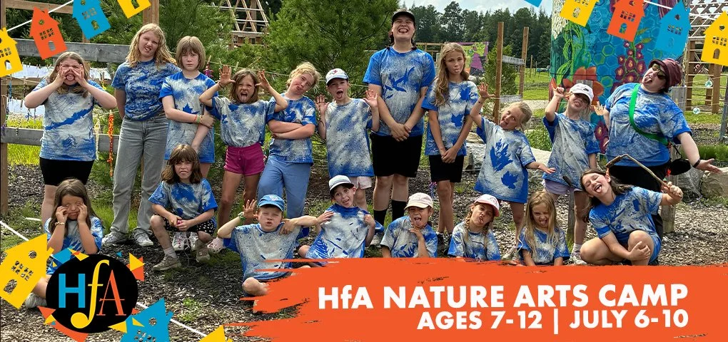 Group of children and a few adults at a nature arts camp, wearing matching tie-dye shirts, outdoors with trees and colorful banners, some making funny faces and poses.