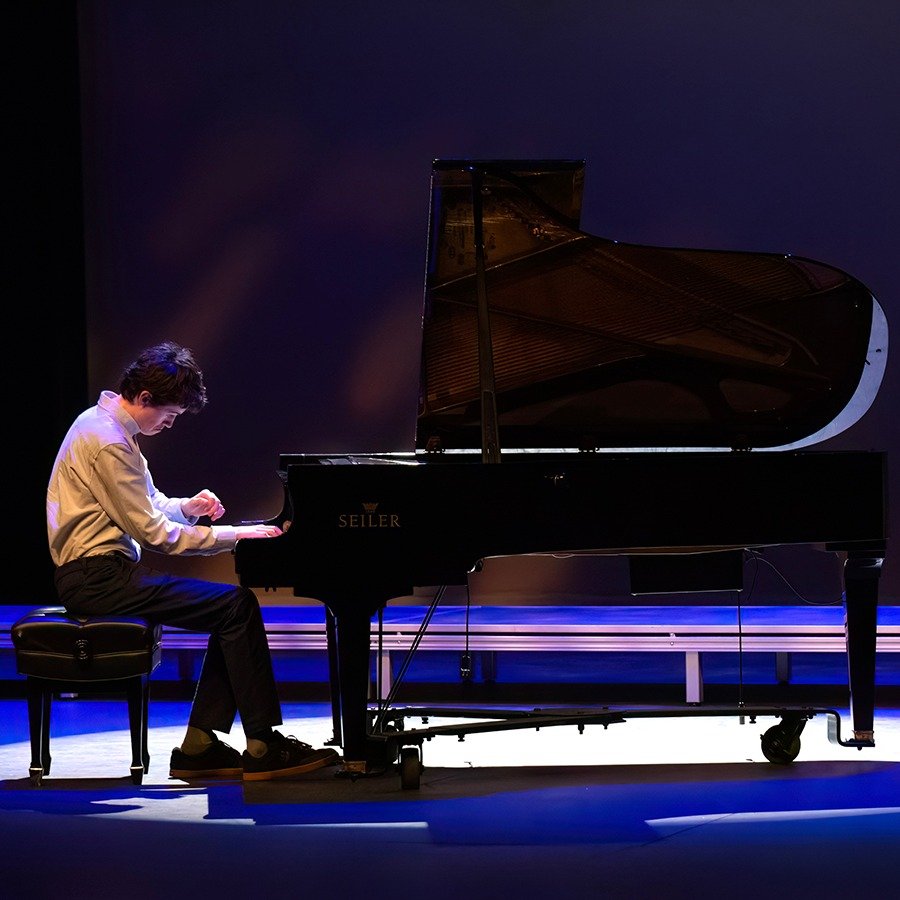 A person playing a grand piano on a stage with purple lighting.
