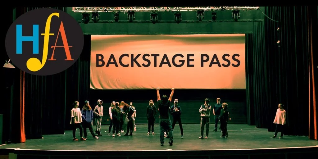 A group of people rehearsing on a stage with a backstage pass sign in the background.