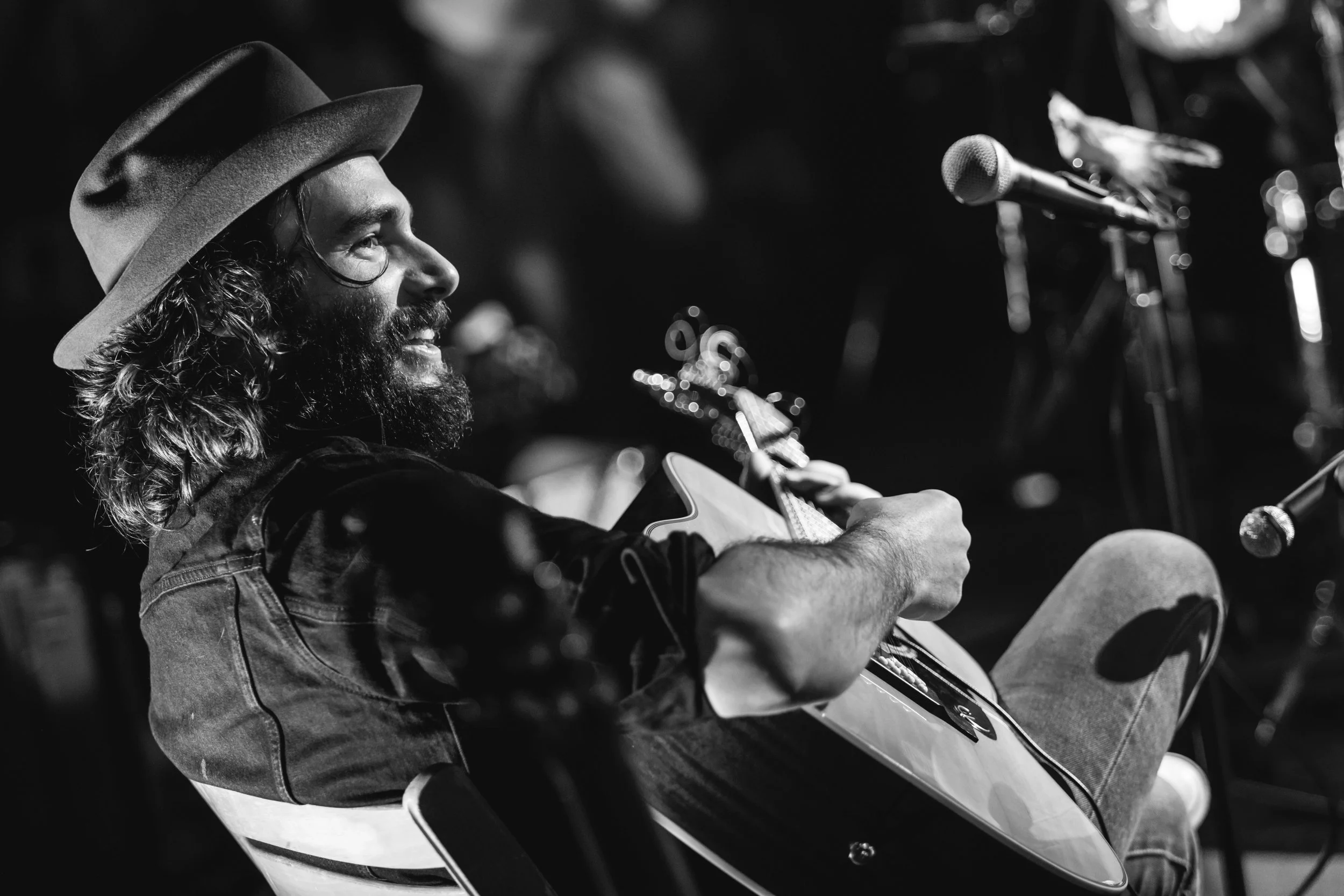 Man with long hair, beard, wearing hat, sitting and playing guitar in black and white, smiling, with microphones nearby.